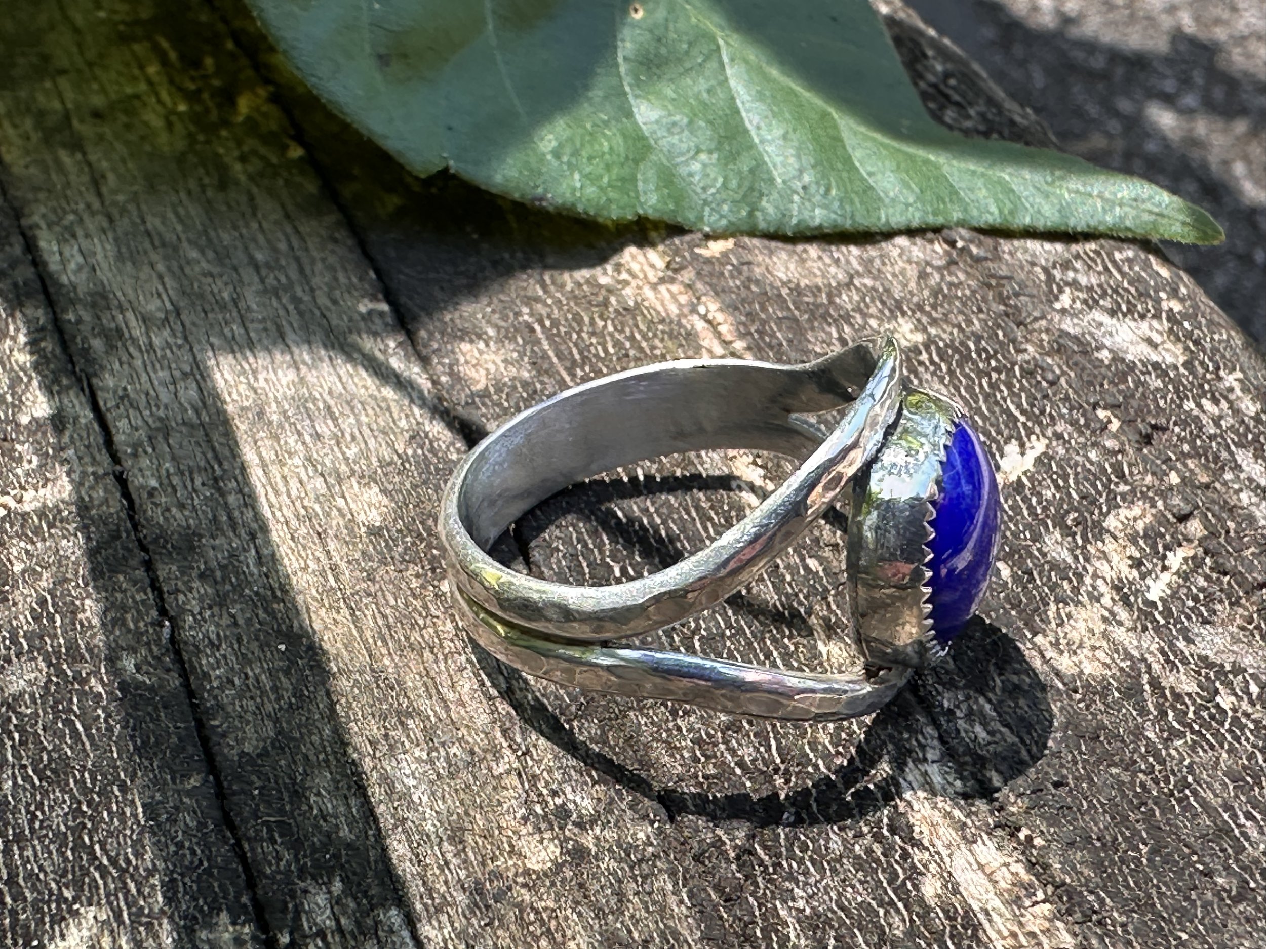 Handcrafted lapis lazuli ring with a deep blue stone in a serrated bezel on a hammered sterling silver split band, resting on weathered wood.