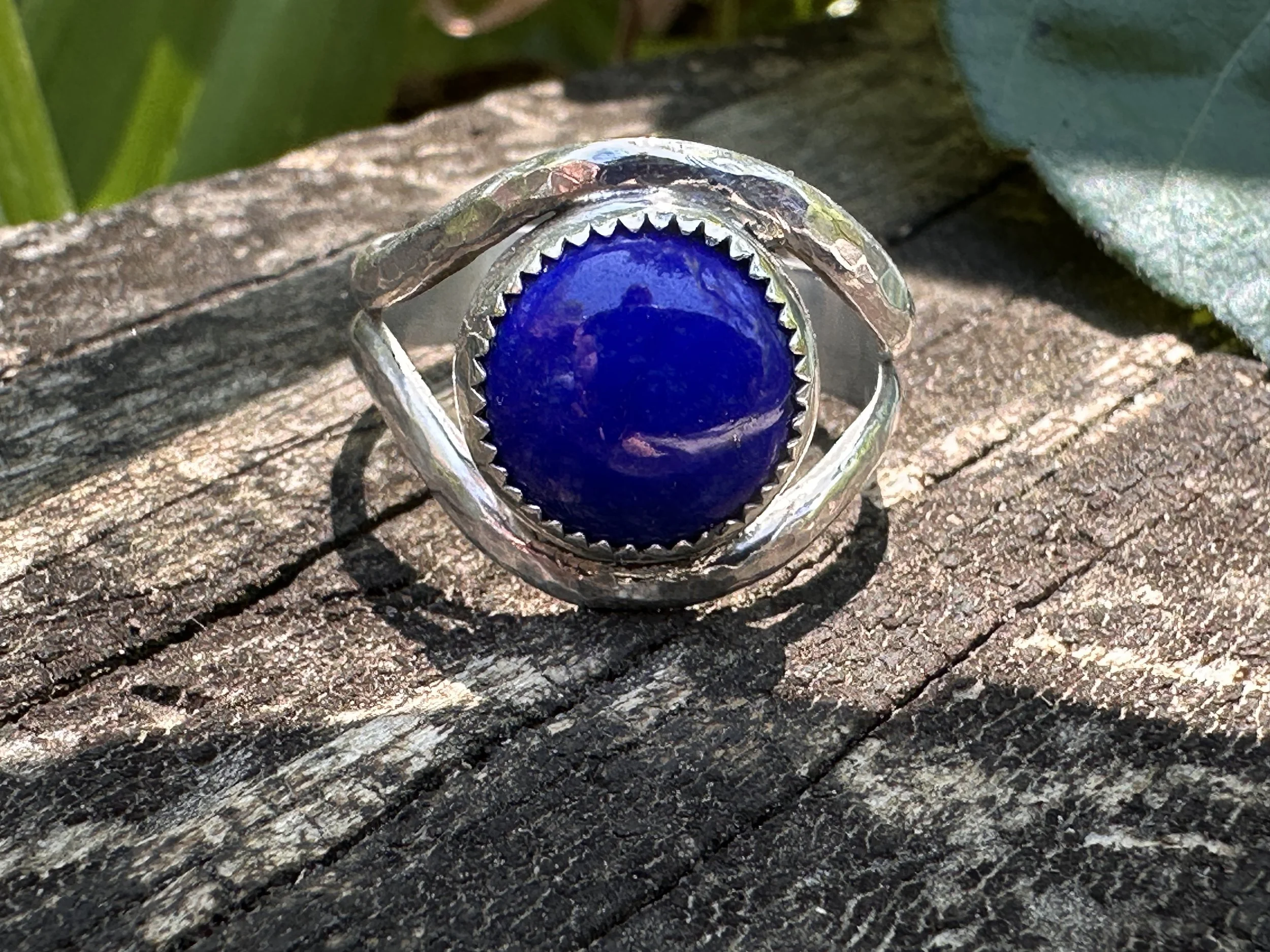 Handcrafted lapis lazuli ring with a deep blue stone in a serrated bezel on a hammered sterling silver split band, resting on weathered wood.