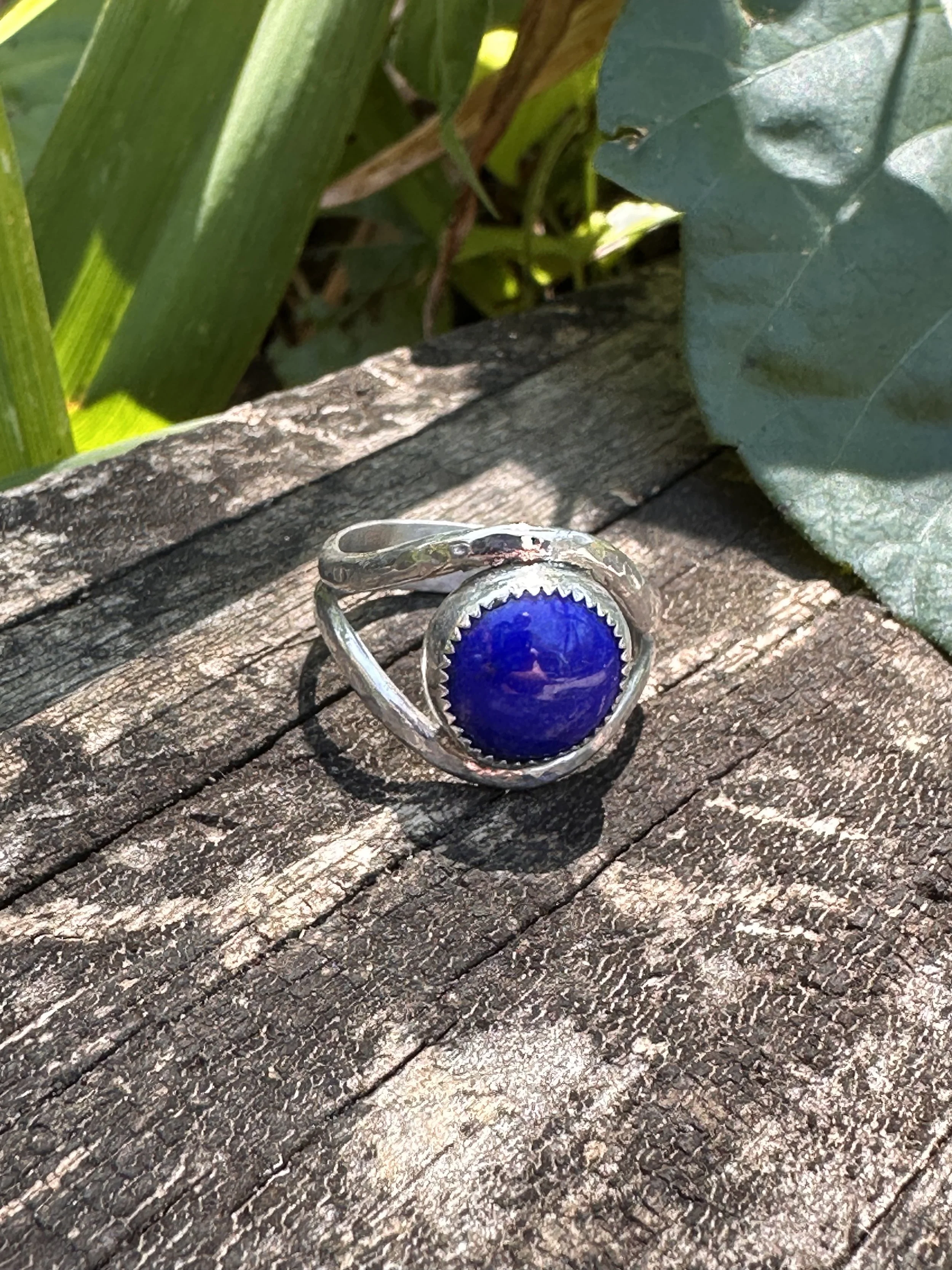 Handcrafted lapis lazuli ring with a deep blue stone in a serrated bezel on a hammered sterling silver split band, resting on weathered wood.