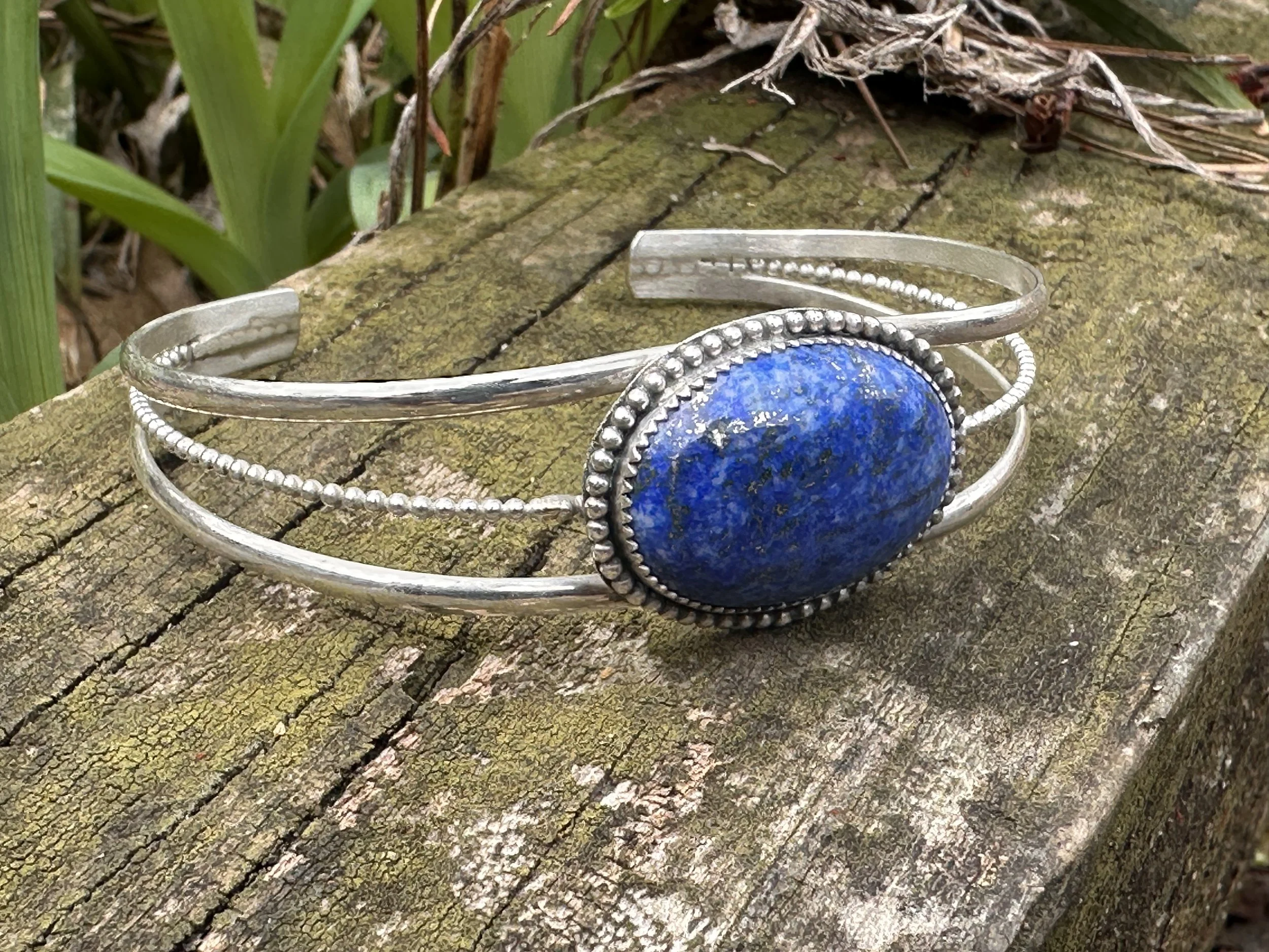 Silver bracelet with large Denim Lapis blue gemstone, resting on a weathered wooden surface outdoors.