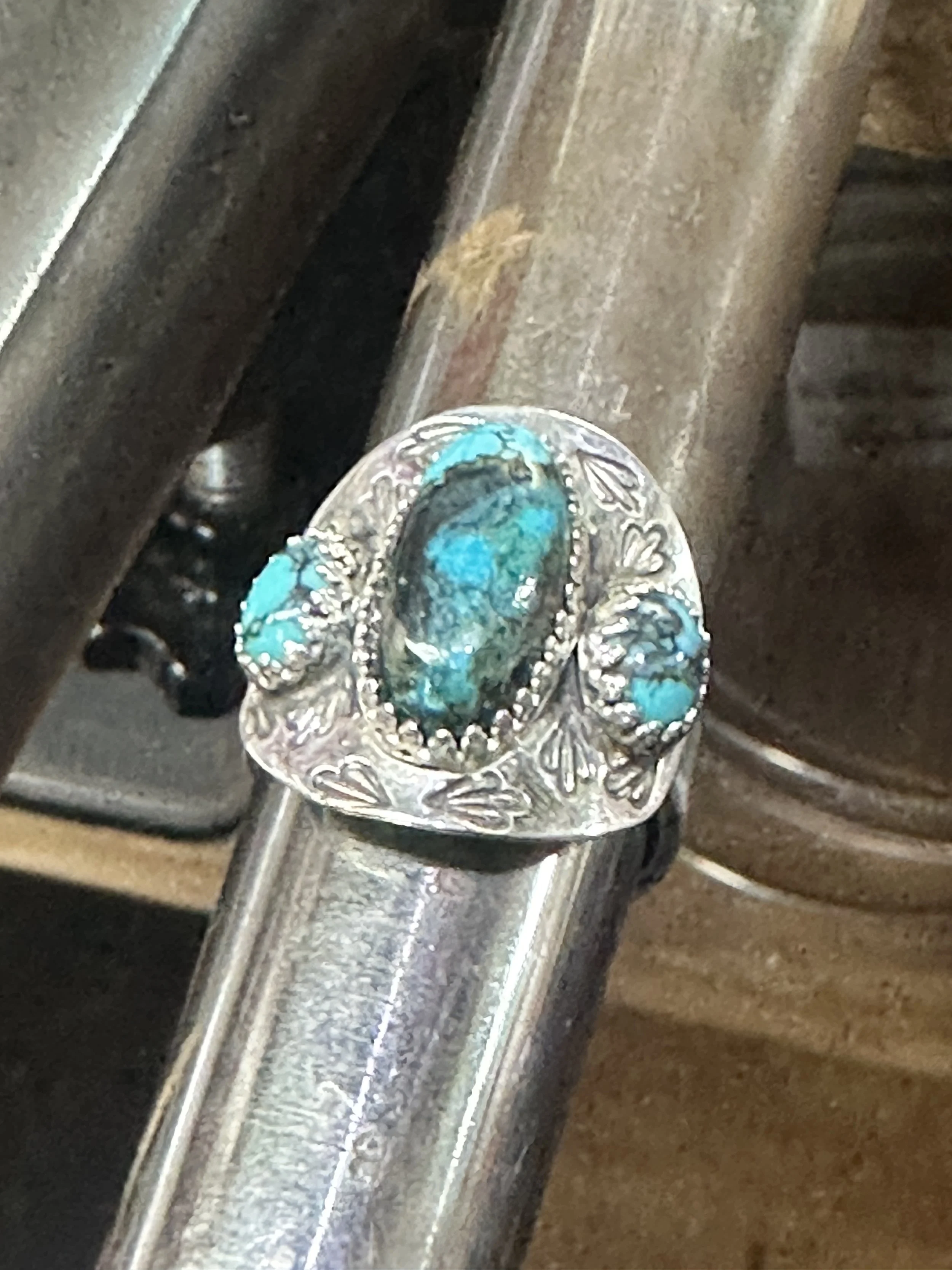 A decorative ring with turquoise stones and intricate silver detailing, resting on a metallic surface.