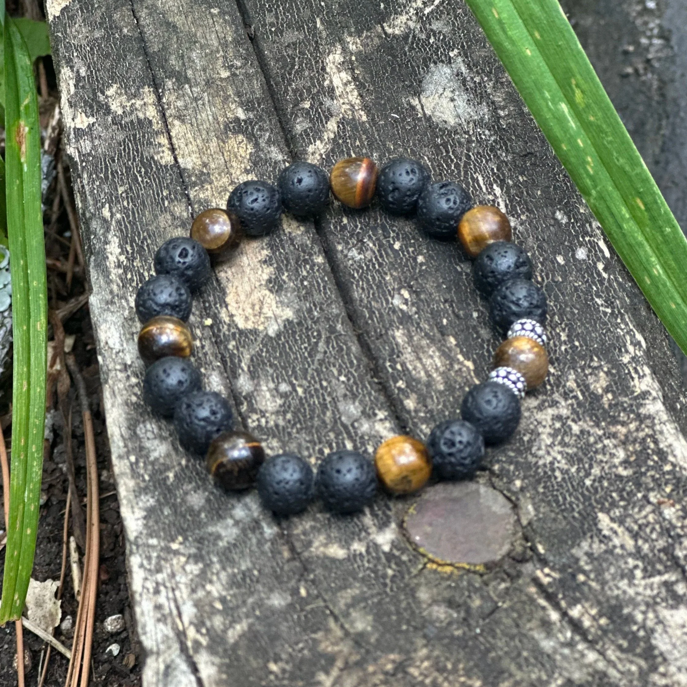 A string of mixed black and brown beads with a few white beads, resting on a weathered wooden surface with green grass blades on either side.