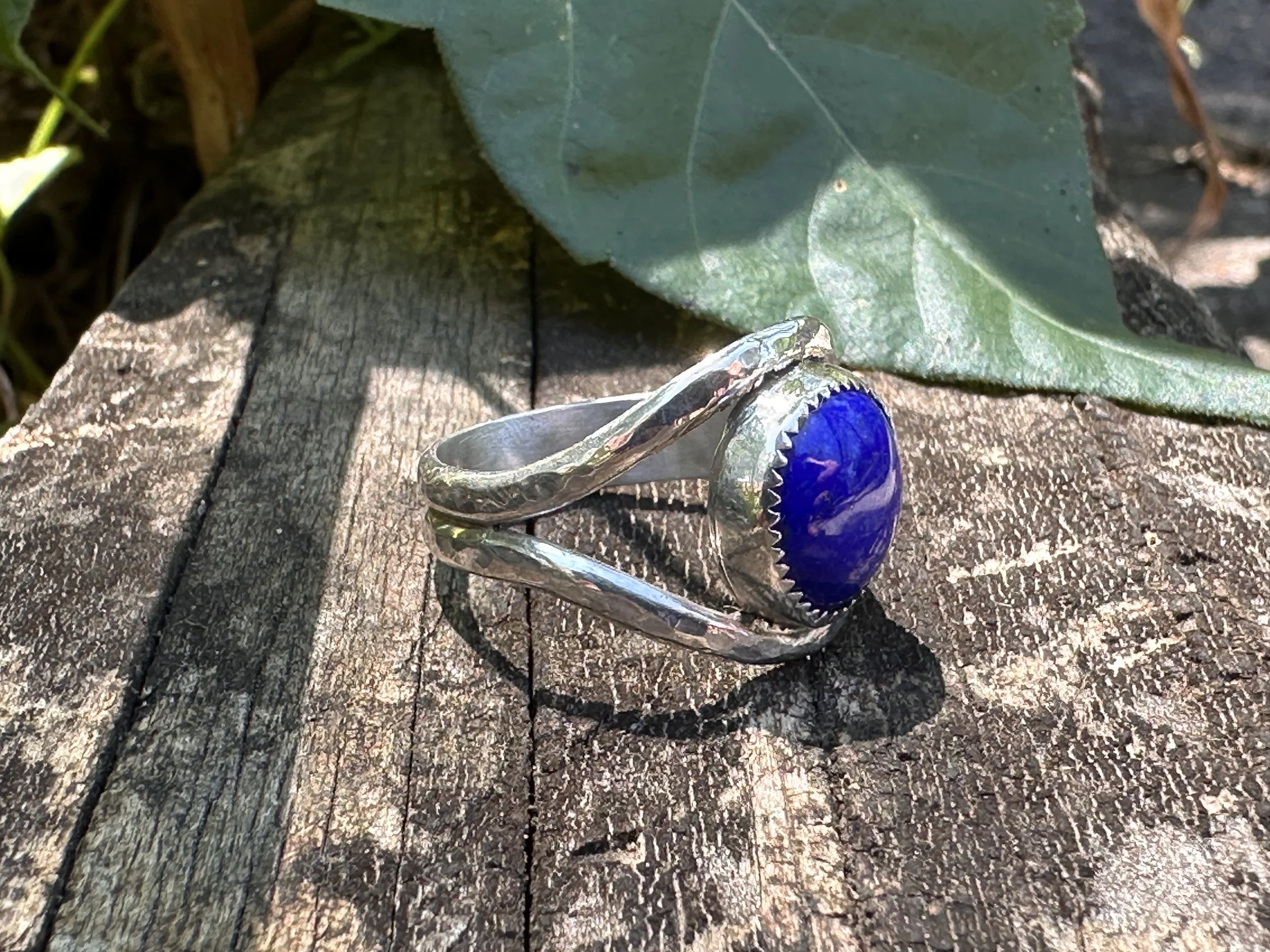 Handcrafted lapis lazuli ring with a deep blue stone in a serrated bezel on a hammered sterling silver split band, resting on weathered wood.