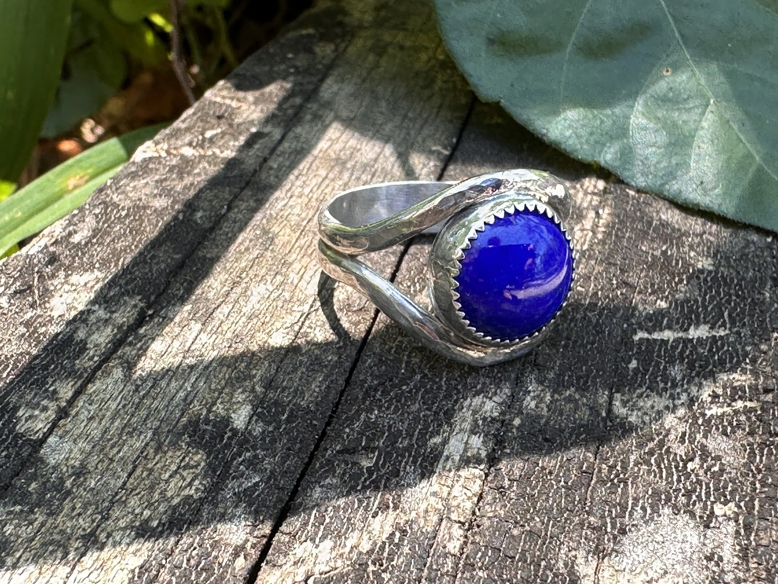 Handcrafted lapis lazuli ring with a deep blue stone in a serrated bezel on a hammered sterling silver split band, resting on weathered wood.