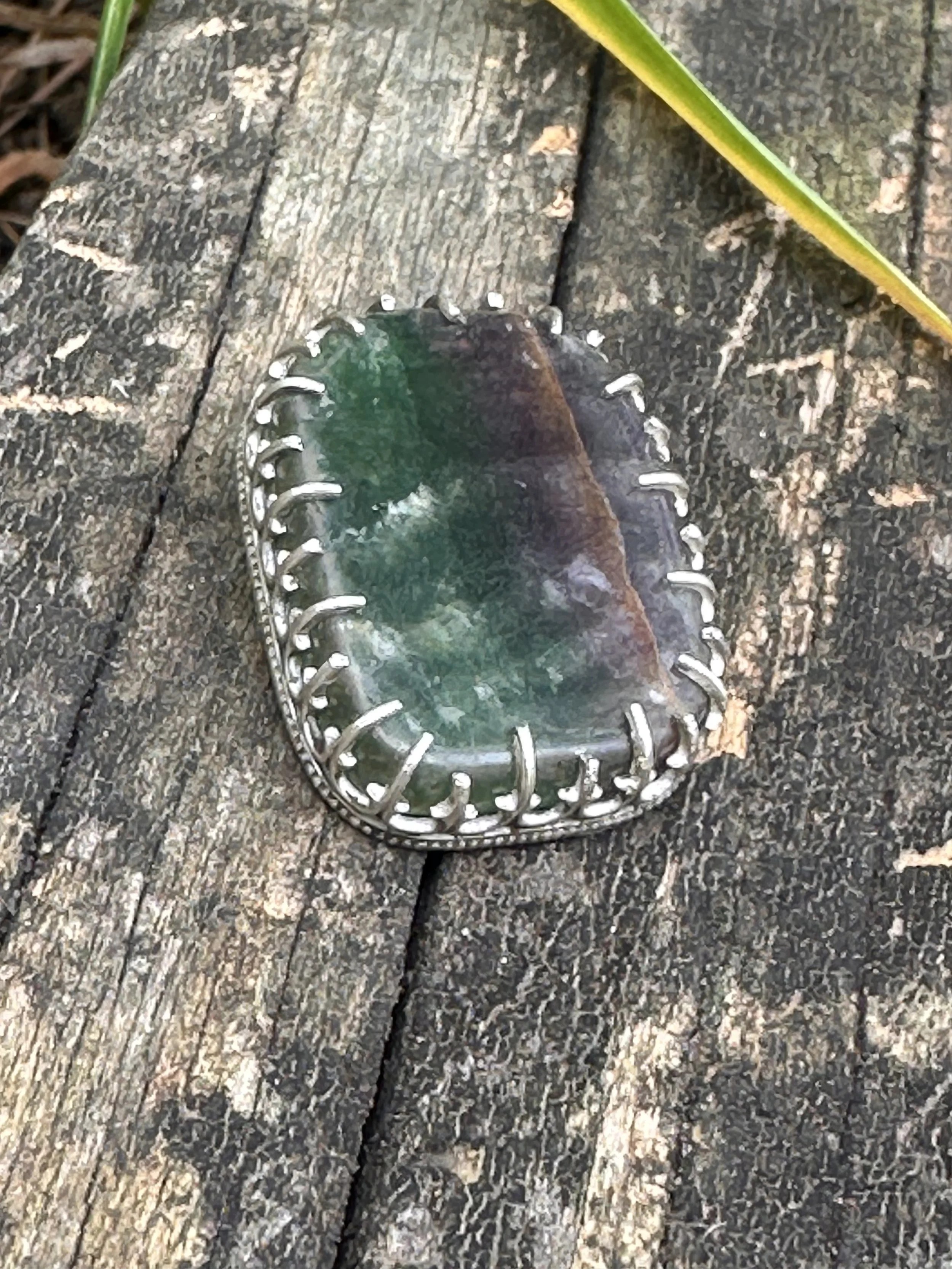 A small, reflective, greenish gemstone set in a silver, decorative pendant with a stitched border, resting on a weathered wooden surface with visible grain and cracks, partially covered by a green leaf.