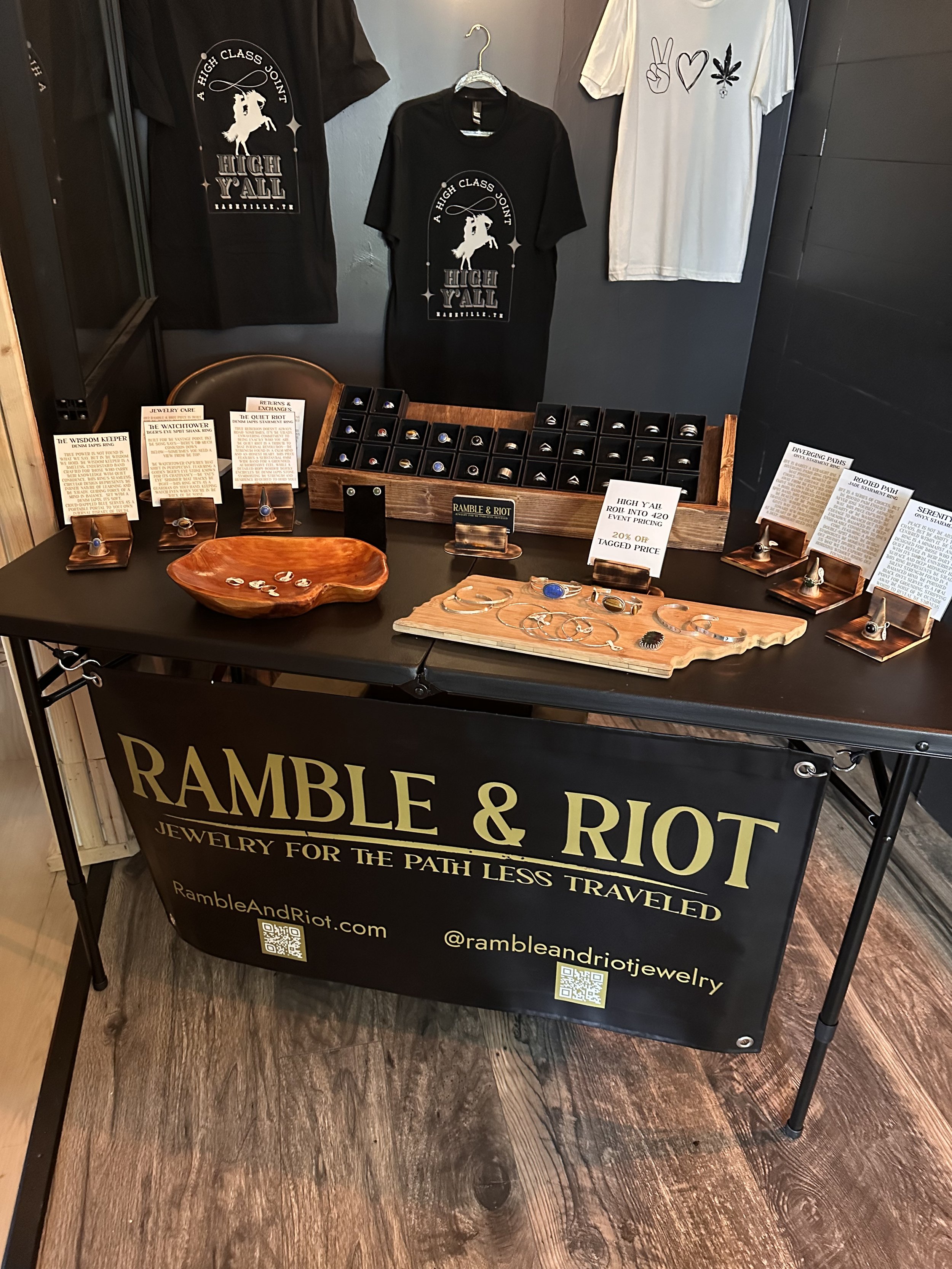 Display table for Ramble & Riot jewelry with rings and rings on stands, promotional signs highlighting discounts, t-shirts with peace symbols, and jewelry in display cases.
