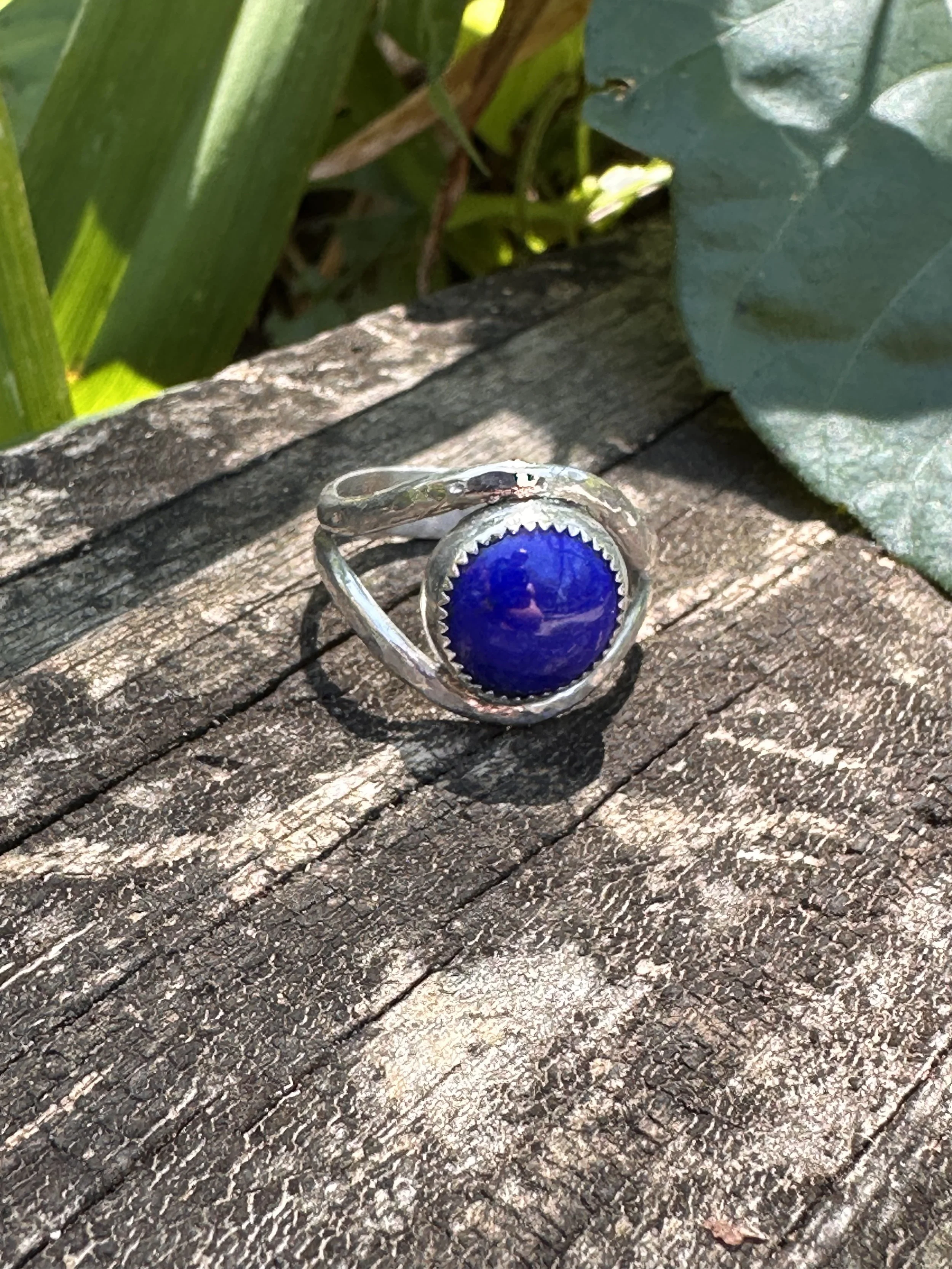 Handcrafted lapis lazuli ring with a deep blue stone in a serrated bezel on a hammered sterling silver split band, resting on weathered wood.