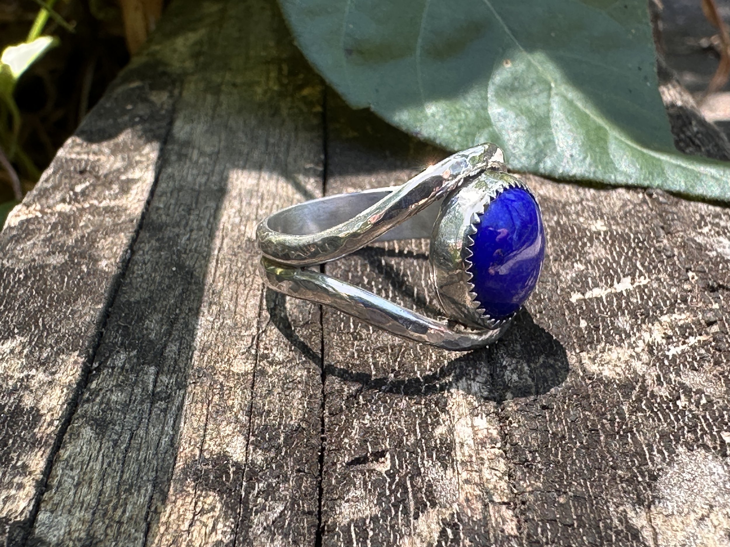 Handcrafted lapis lazuli ring with a deep blue stone in a serrated bezel on a hammered sterling silver split band, resting on weathered wood.