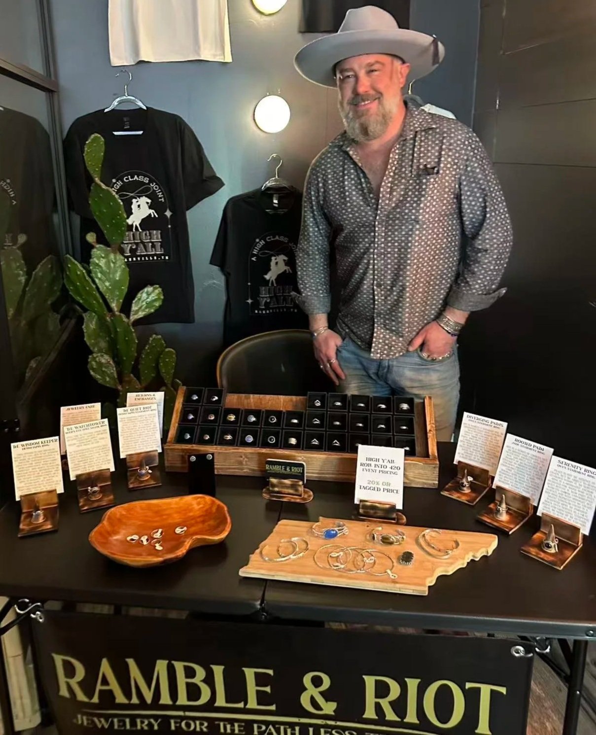 A man wearing a large gray hat and patterned shirt stands behind a jewelry display table at a booth with a sign that reads "Ramble & Riot Jewelry for the Path Less Traveled." On the table are various jewelry pieces, informational cards, and a wooden tray with rings. There is a large cactus plant to the left of the table.