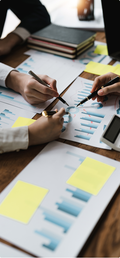 People are working on financial charts and graphs at a desk, with notebooks, sticky notes, and a calculator.