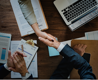 Two people shaking hands over a desk with documents, a laptop, and notebooks.