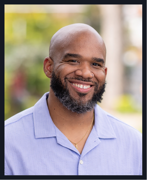 Portrait of a smiling Black man with a bald head and beard, wearing a light blue collared shirt, outdoors with blurred greenery in the background.