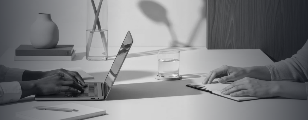 Two people sitting at a desk, one typing on a laptop and the other writing in a notebook, with a glass of water, a pen, and some decorative items on the desk.