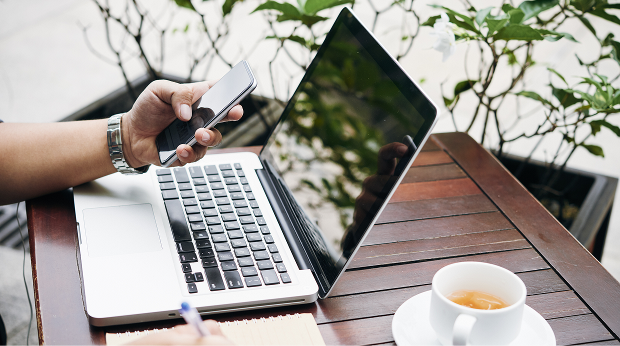 A person using a smartphone while sitting at a wooden table with an open laptop, a notebook, a pen, and a cup of tea.
