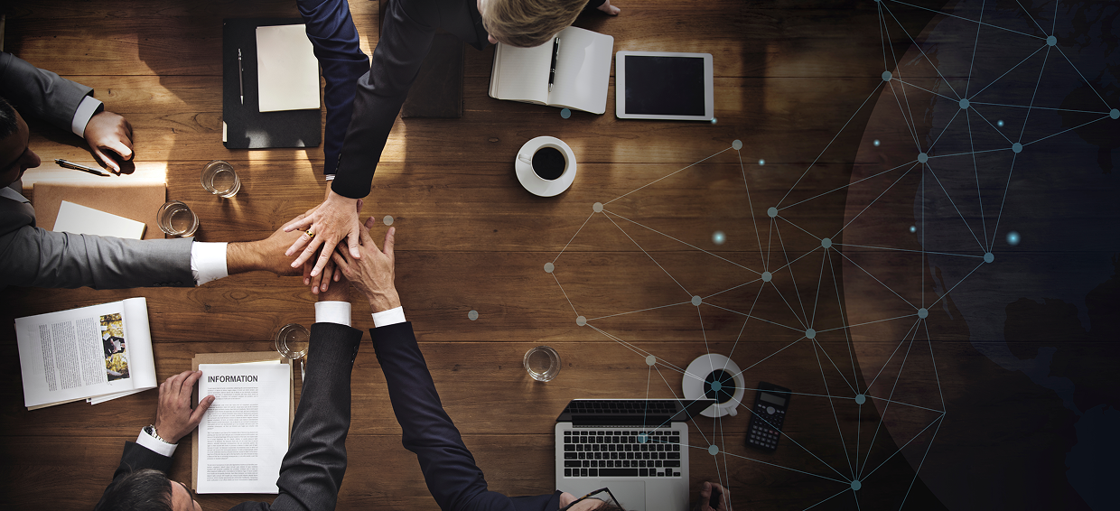 Business people in suits reaching for a handshake over a wooden conference table with laptops, notebooks, cups of coffee, and a tablet.