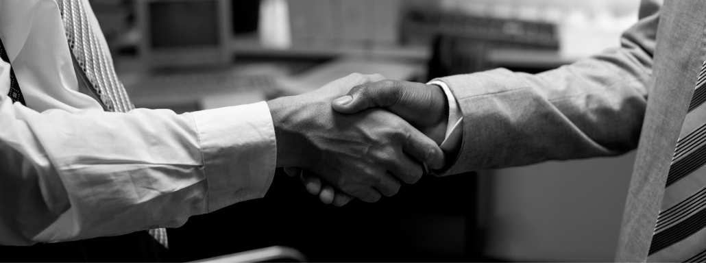 Close-up of two business people shaking hands in an office setting, one in a light-colored suit and the other in a checked shirt and tie.