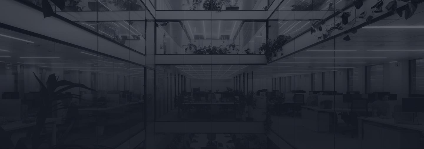 A modern office with glass walls, workstations, and potted plants, viewed from a hallway.