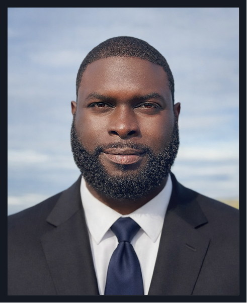 Portrait of a man wearing a dark suit, white shirt, and navy tie, outdoors with a cloudy sky background.