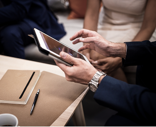 Person using a tablet with other people sitting nearby in a meeting or casual setting.