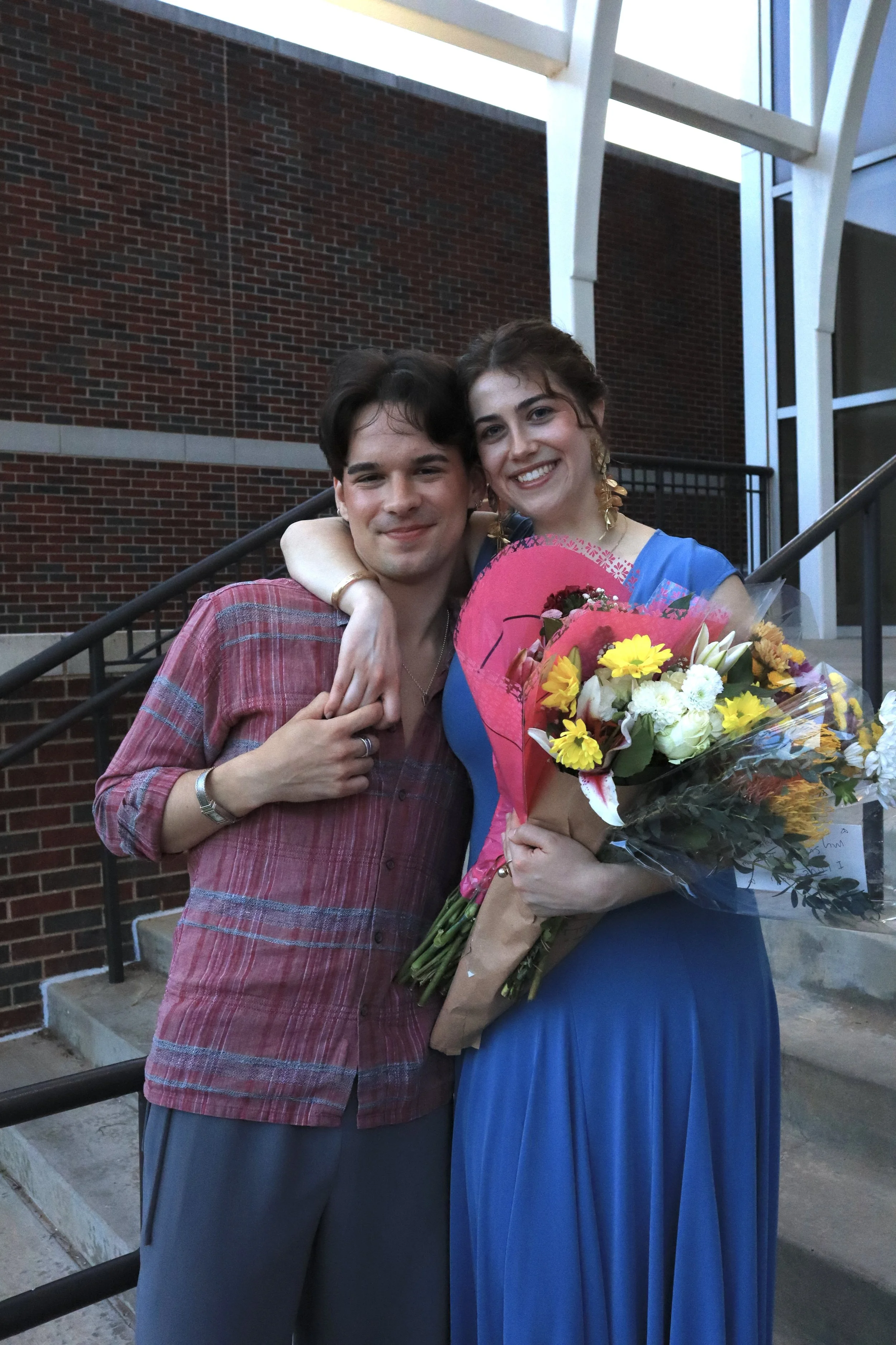 A young man and woman are smiling and hugging each other, the woman holding a bouquet of colorful flowers, in front of a brick building.