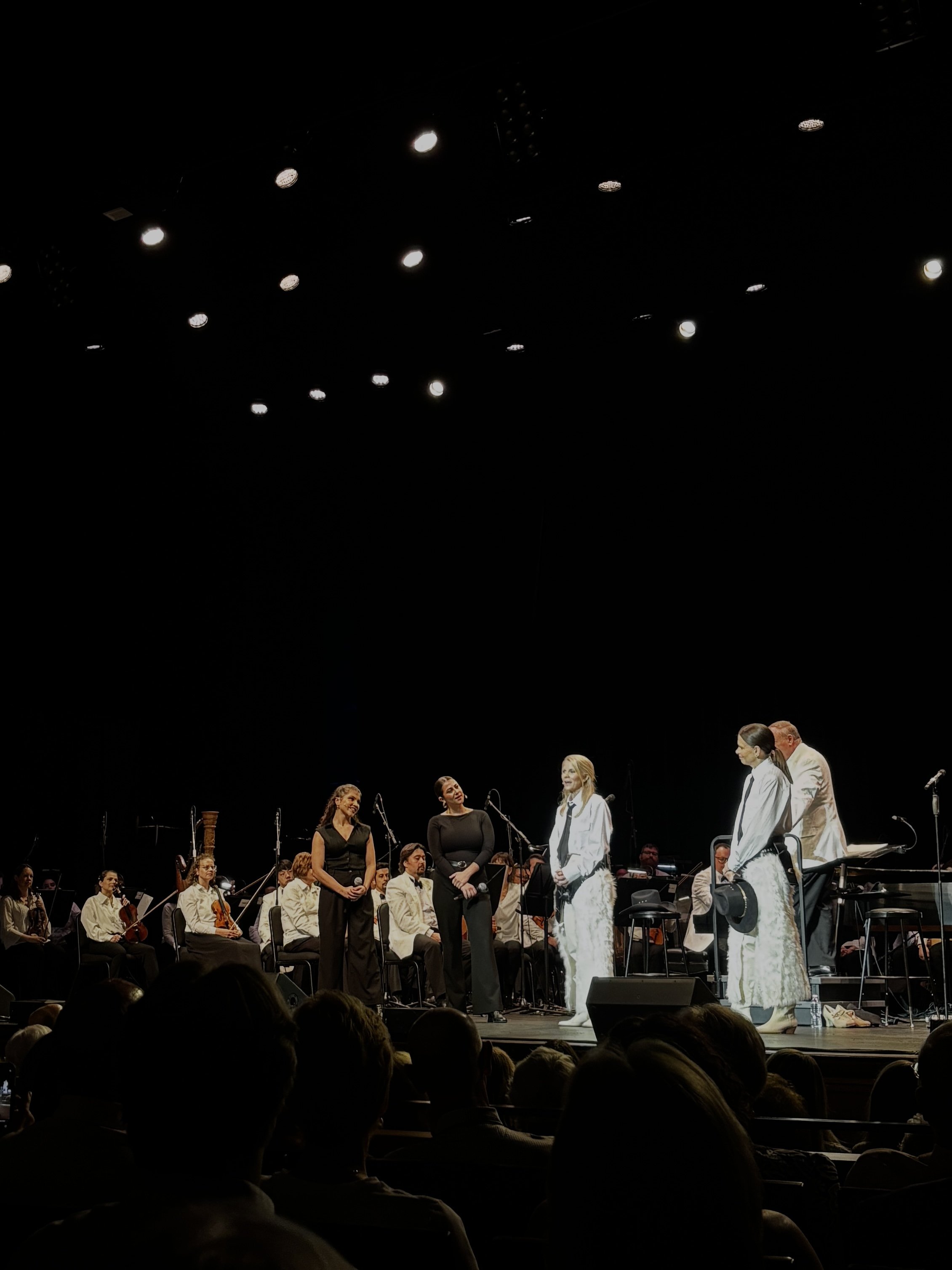 A group of musicians and singers on stage during a concert or performance, with an audience in the foreground. The stage is lit with bright overhead lights, and the performers are dressed in formal or semi-formal attire.