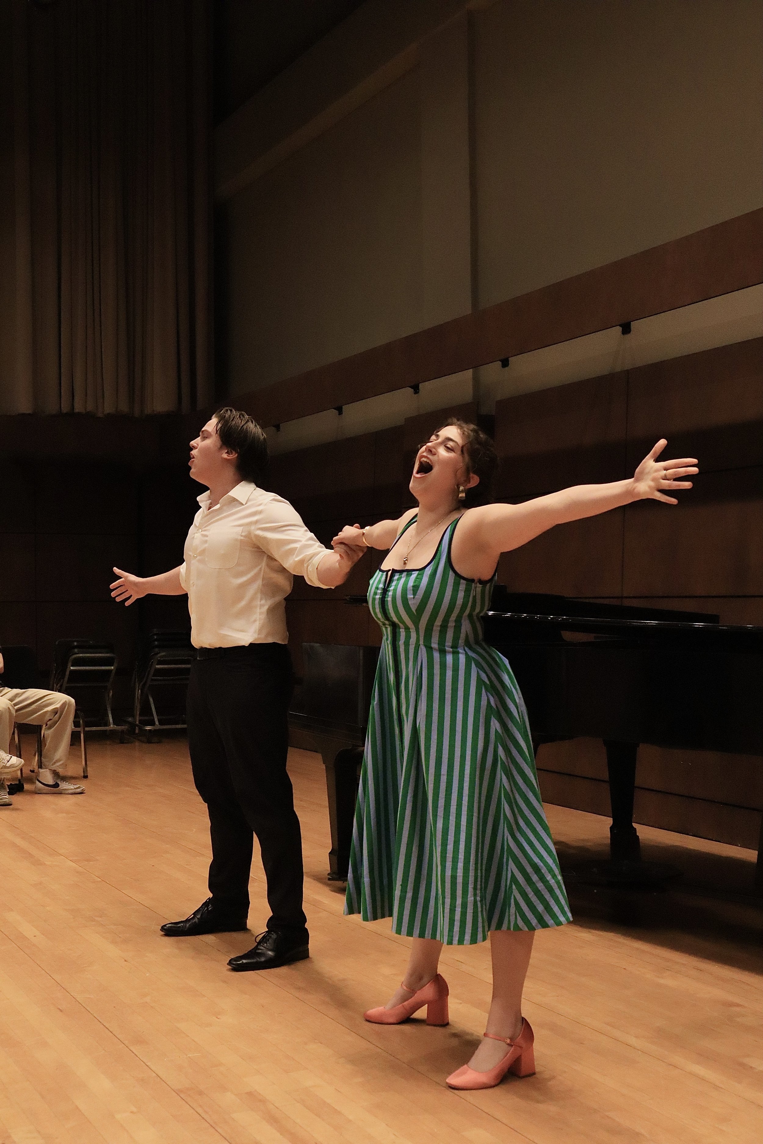 Two performers on stage, one woman with arms wide open, wearing a striped green and white dress with pink heels, and one man with arms outstretched, wearing a beige shirt and black pants, holding hands as if singing or performing.