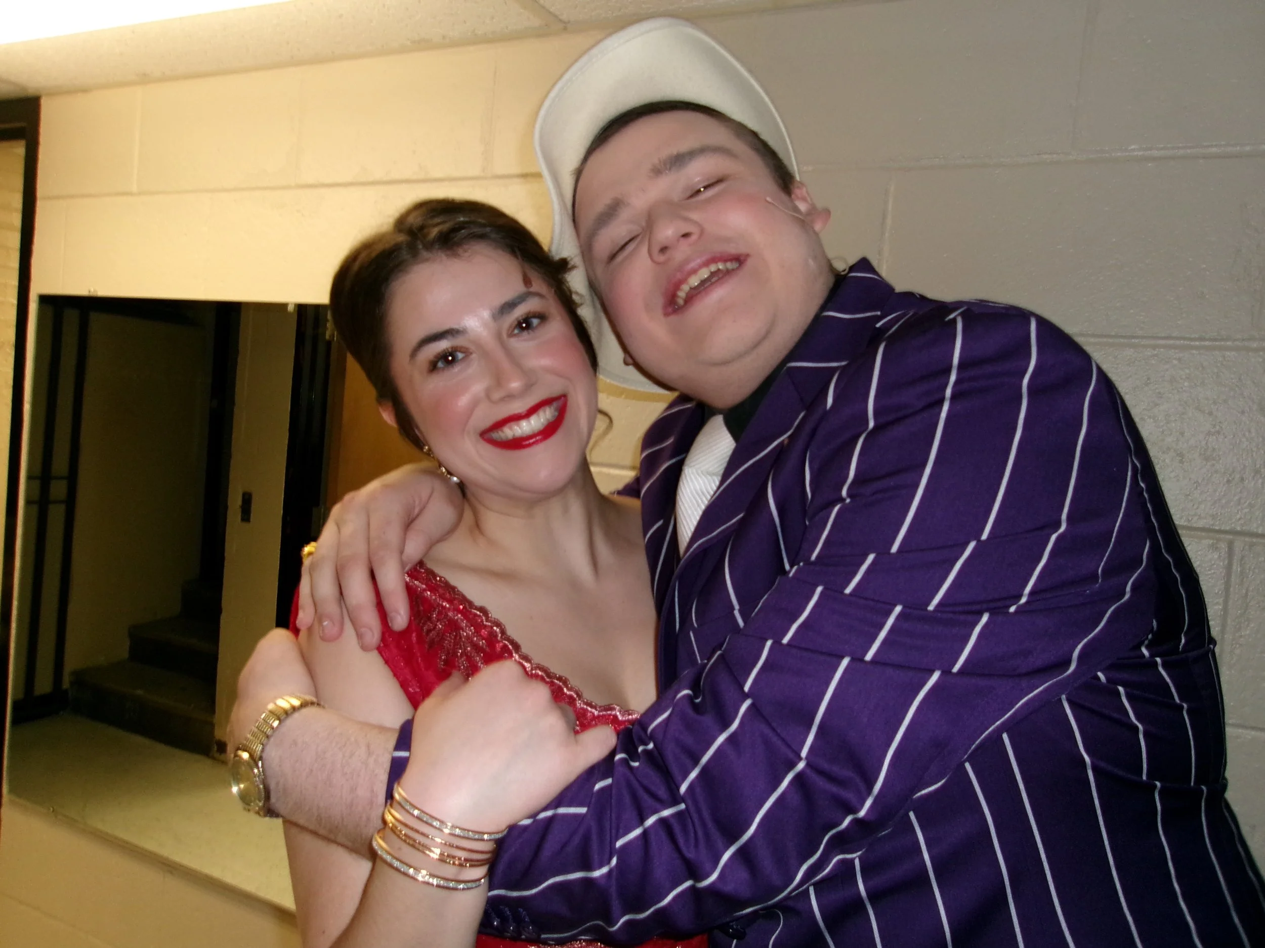 A woman in a red dress and a man in a purple striped suit are embracing and smiling, with the woman resting her head on the man's shoulder.
