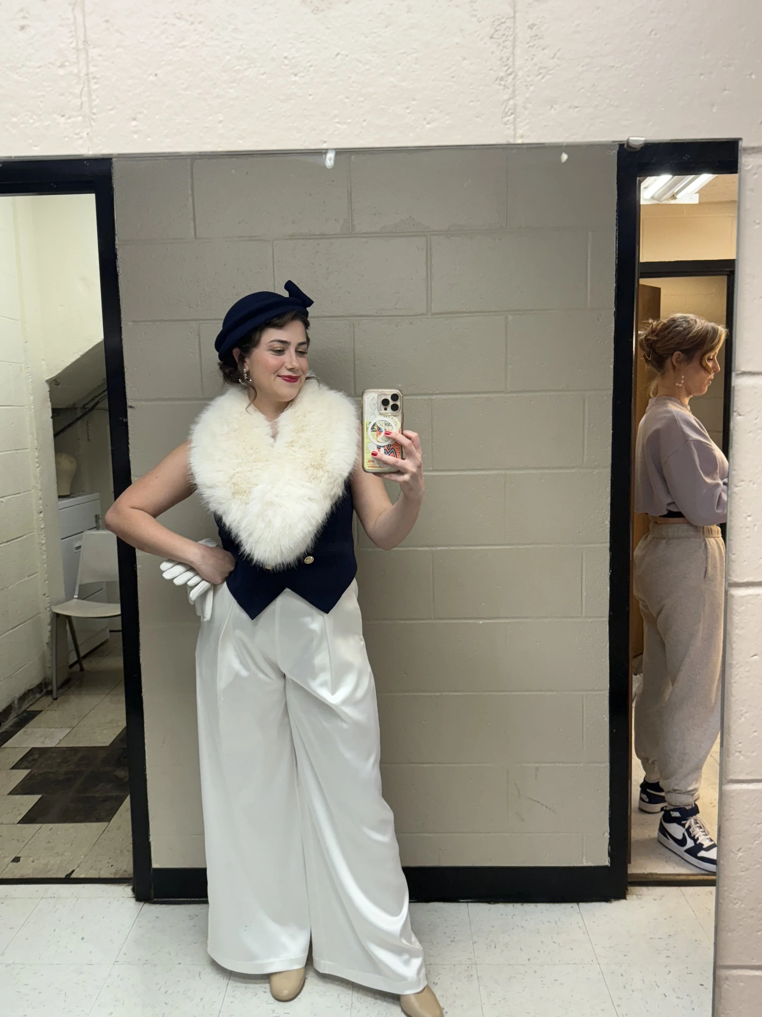 Two women taking a mirror selfie in an indoor setting, one wearing a dark hat, white faux fur stole, and white wide-leg pants, the other wearing beige sweatpants and sneakers, with a beige sweatshirt tied around her waist.