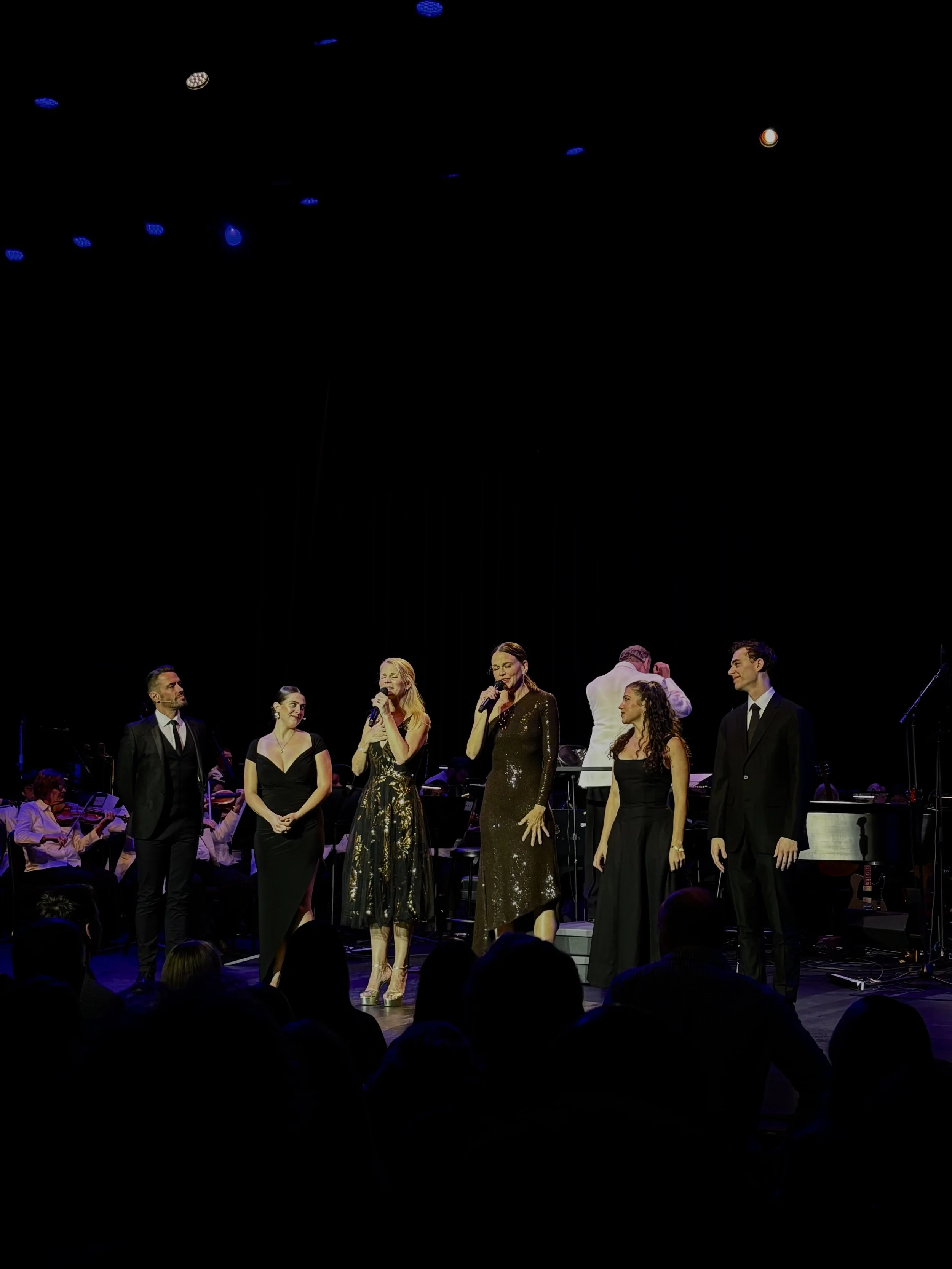 A group of performers on stage during a musical or theatrical performance, with a conductor or singer at the center, dressed in elegant black and gold attire, surrounded by orchestra musicians.