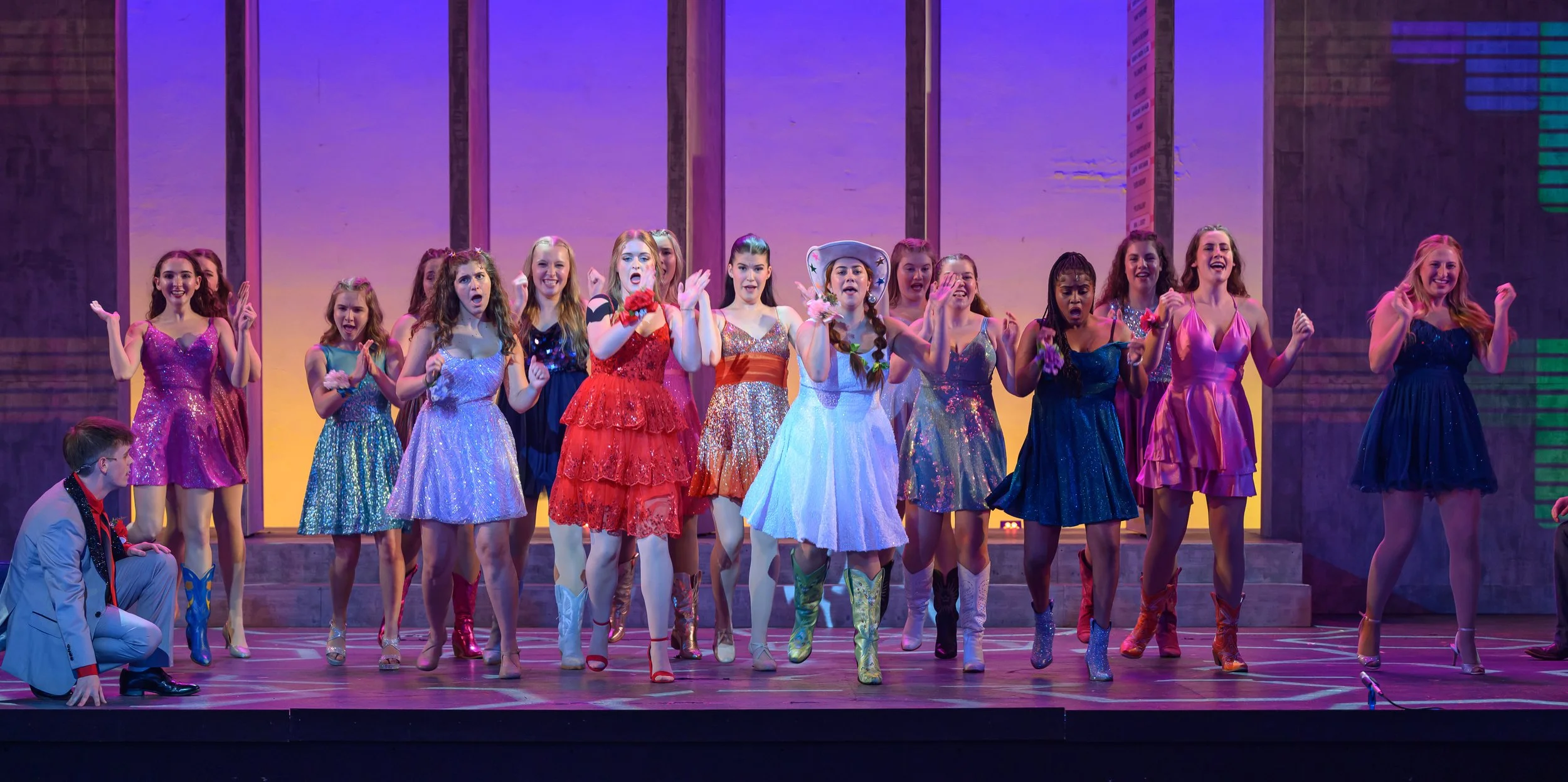 A group of young women and men performing on stage in colorful costumes, with a purple and pink background.