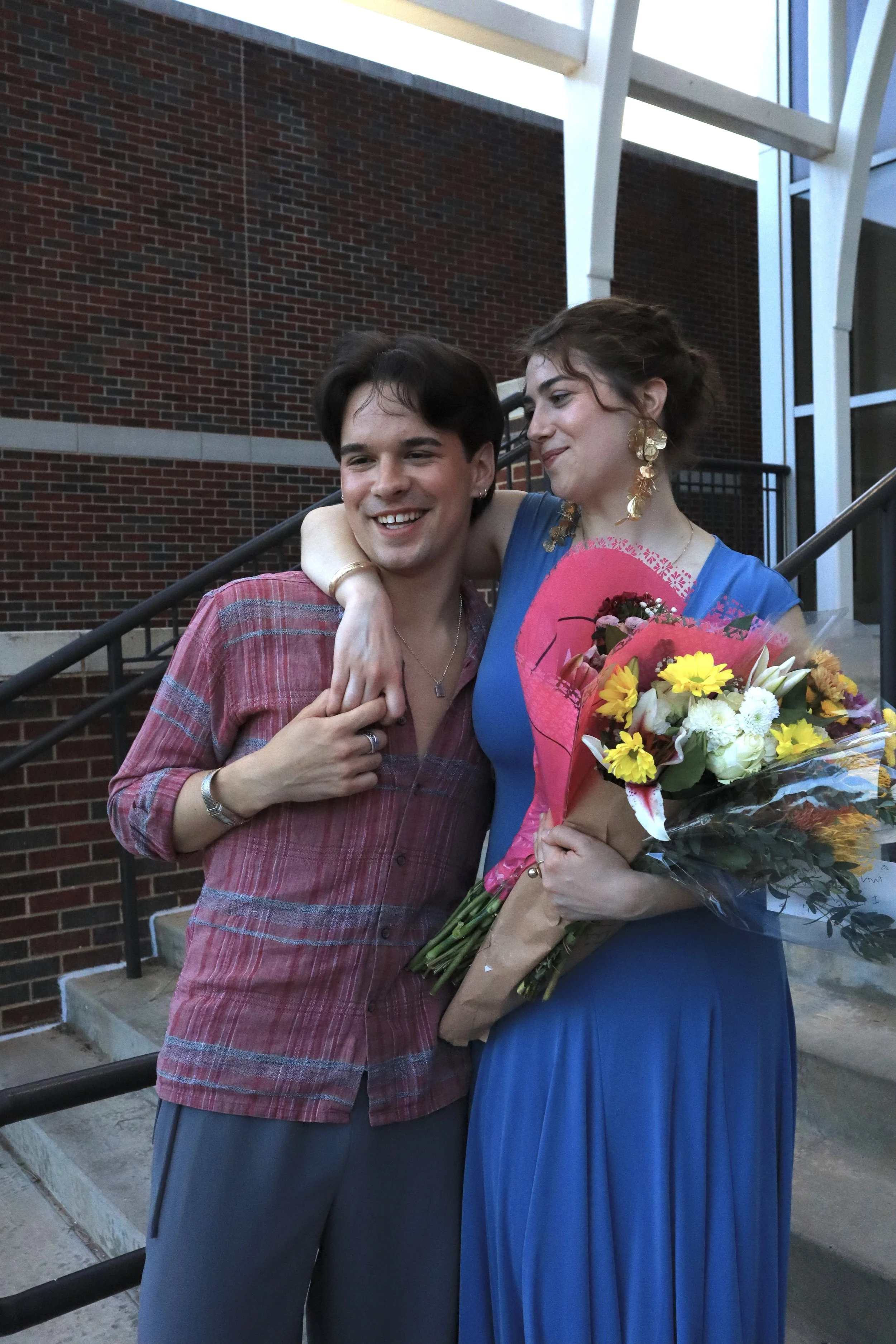 A man and woman celebrating with the woman holding a large bouquet of flowers. The man has short dark hair, wearing a red plaid shirt and smiling, while the woman has curly hair, wearing a blue dress and gold jewelry, and looks fondly at the man. The
