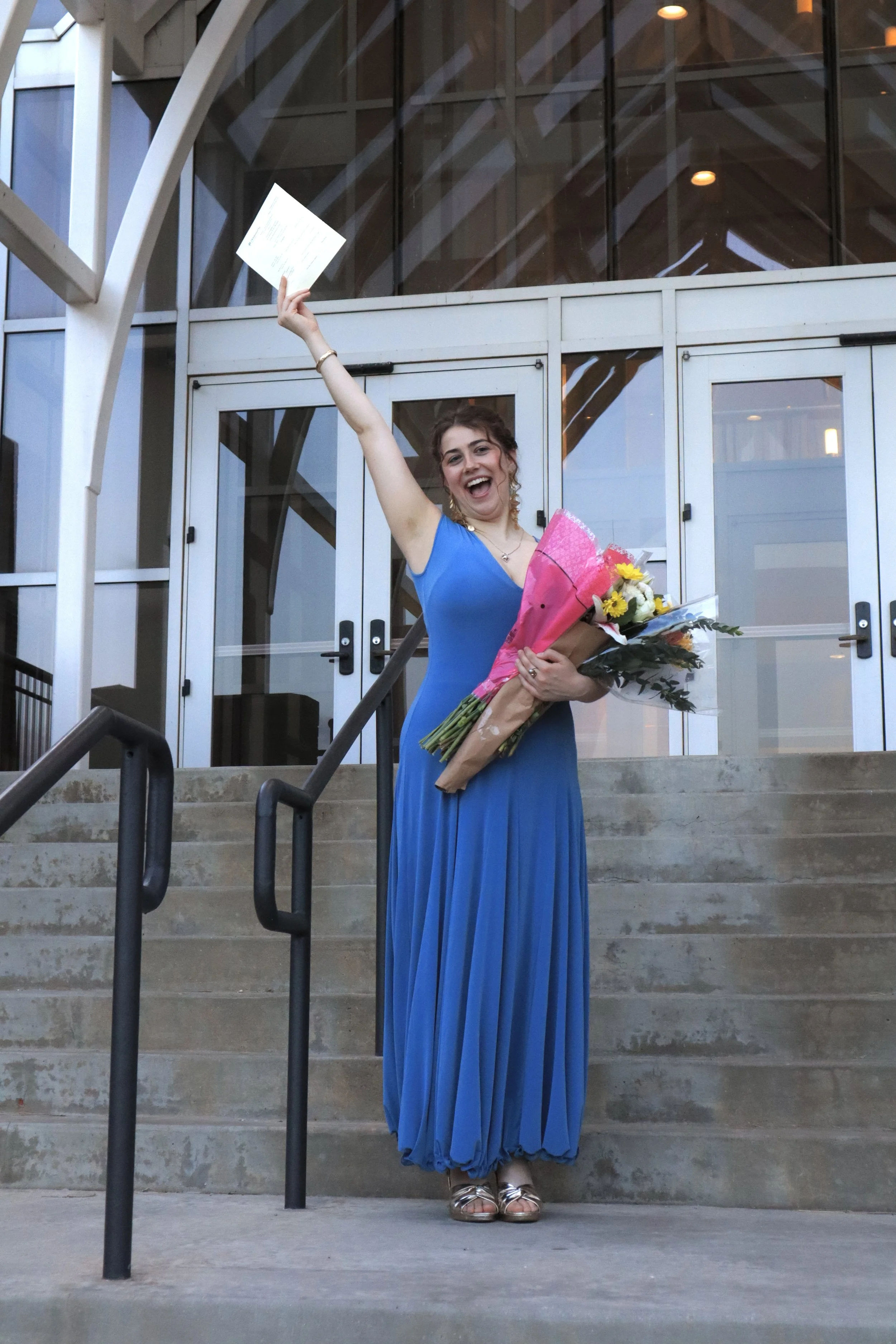 A woman in a long blue dress standing on outdoor stairs, holding a bouquet of flowers in one arm and a document in the other, smiling happily.