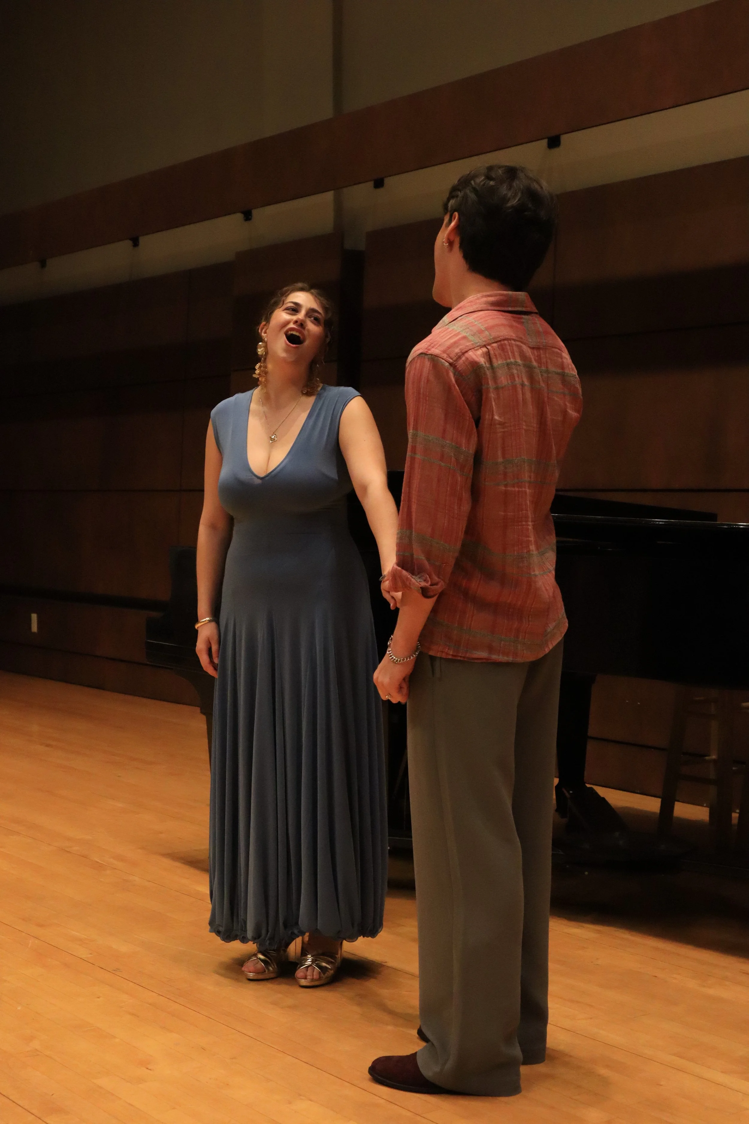 A woman in a long blue dress singing or speaking with her head tilted back, standing next to a man in a plaid shirt and gray pants, on a wooden stage in a room with dark wood paneling.