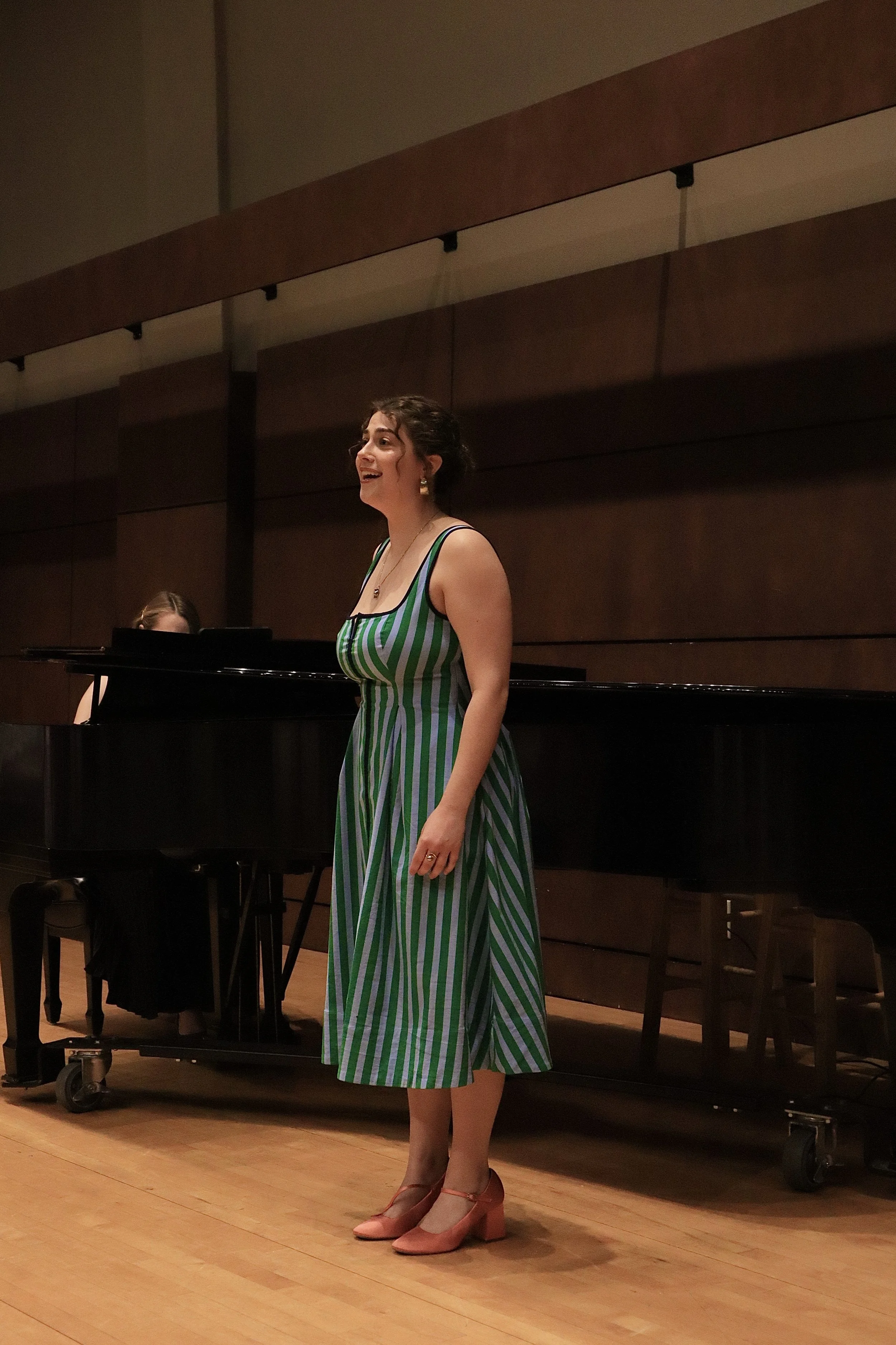A woman in a green and white striped dress singing on stage with a piano and pianist in the background.