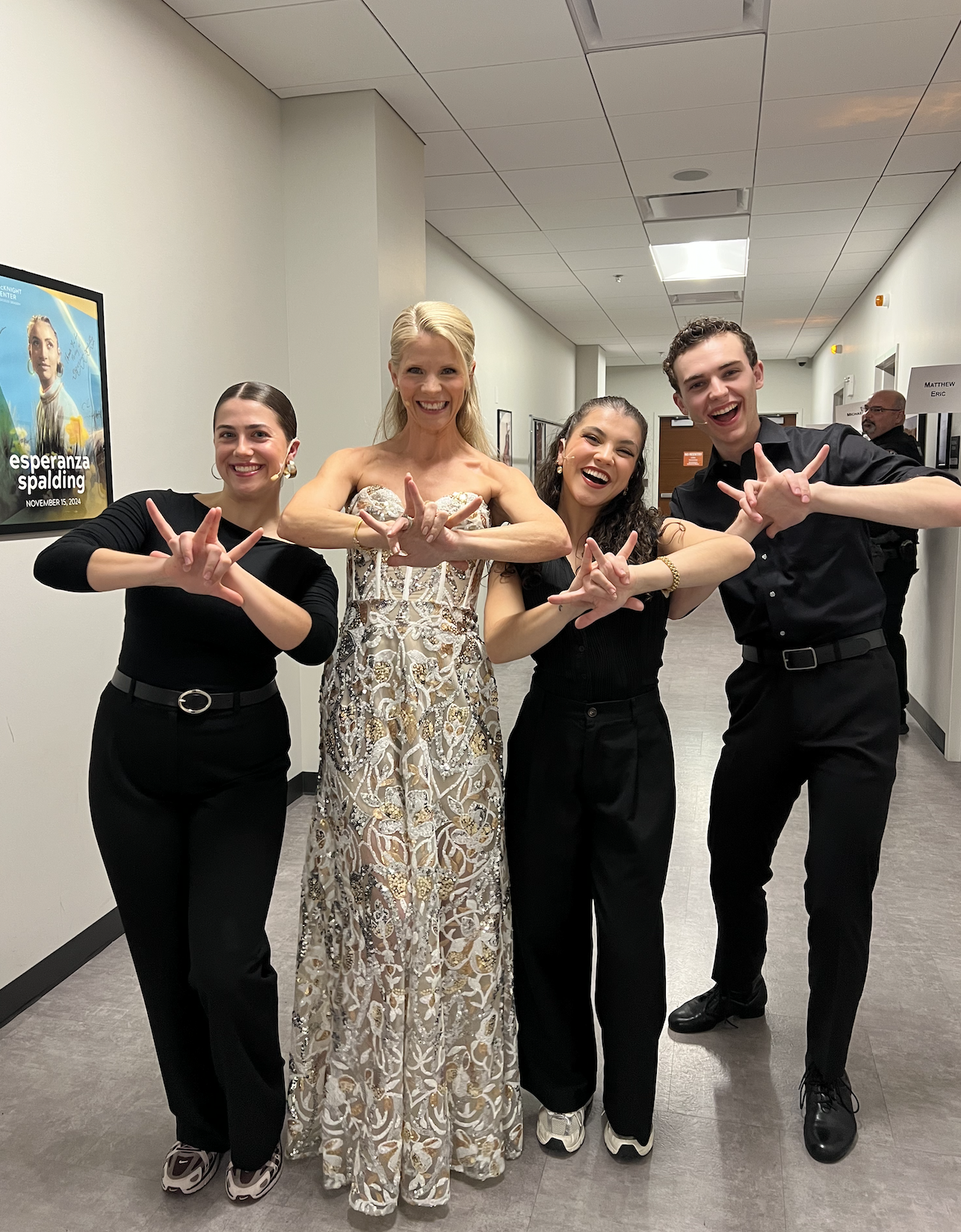 Four people standing in a hallway, making hand gestures, smiling at the camera; one woman is wearing an elegant, strapless, patterned dress, others are in black outfits.