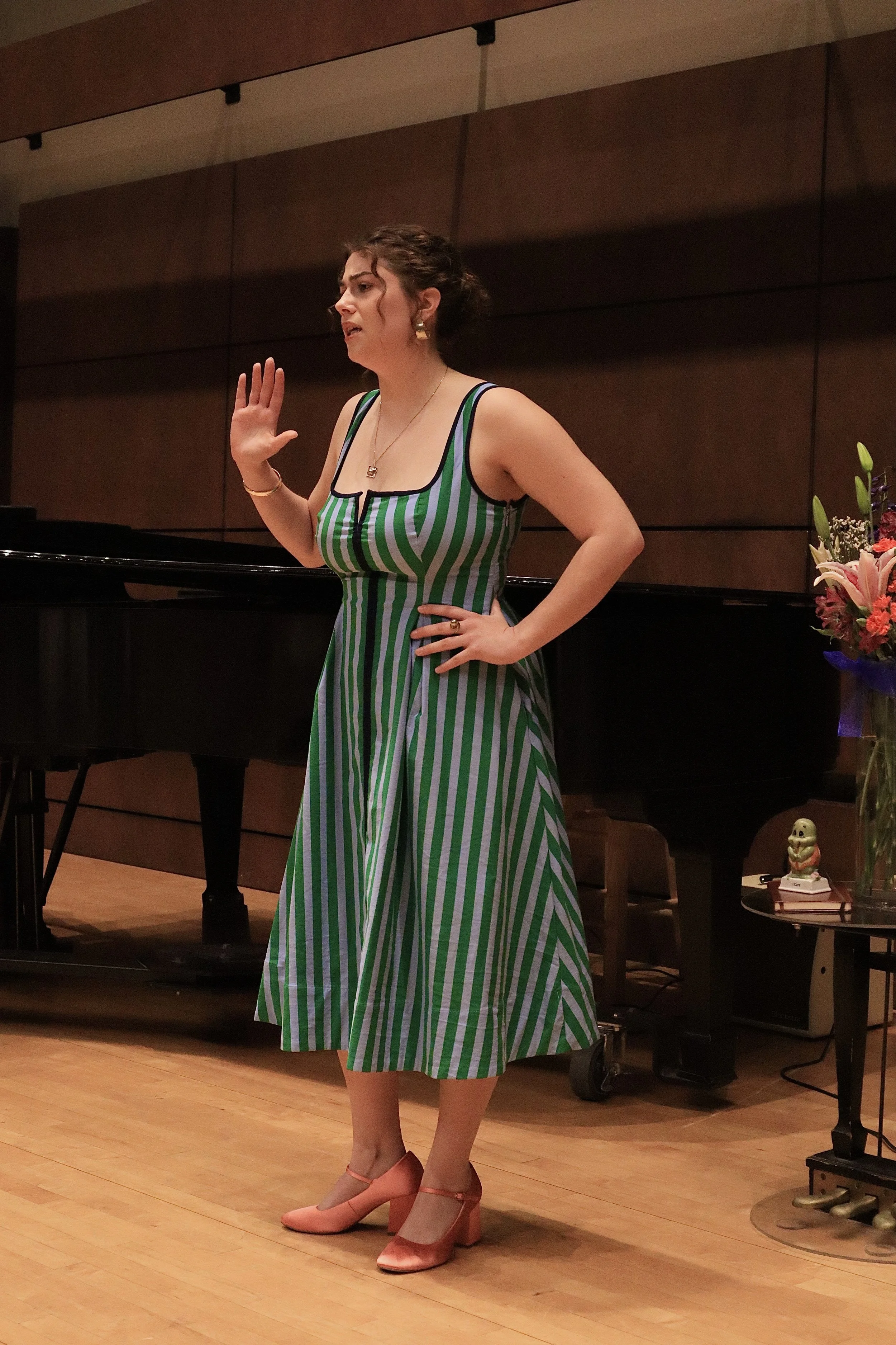 Woman in a green and white striped dress standing on stage, raising her right hand with her palm open while performing in front of a grand piano and a flower arrangement.