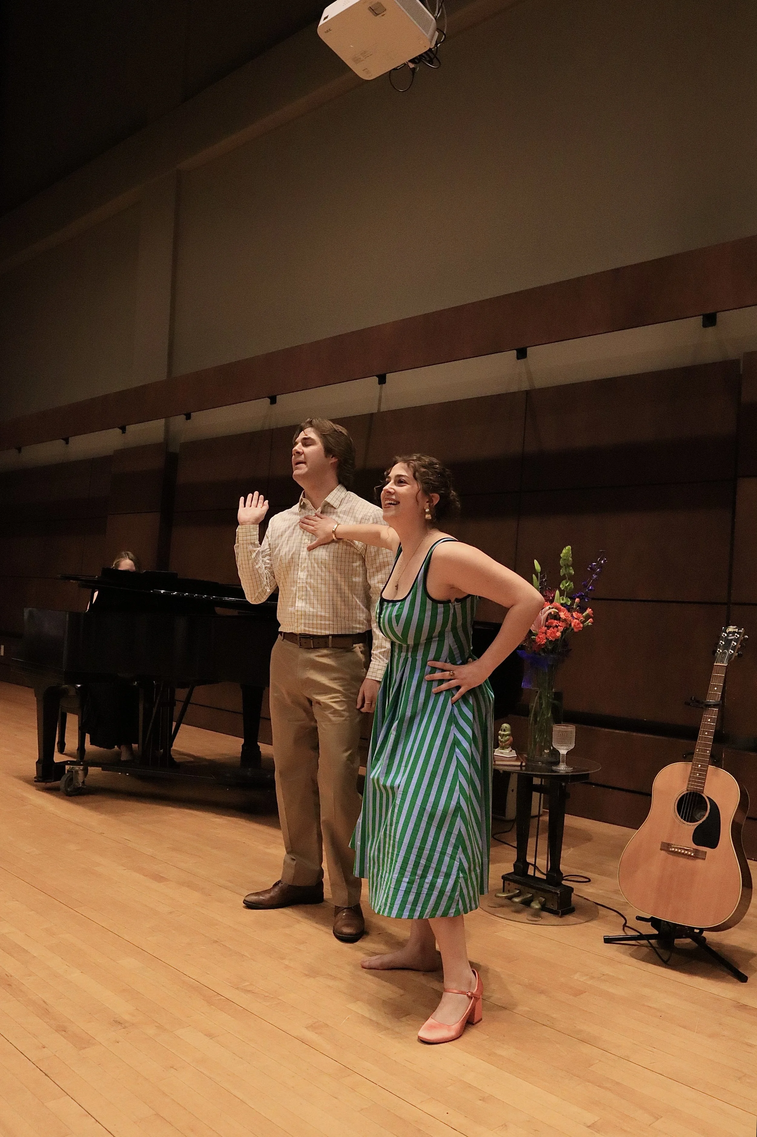 A young woman in a green and white striped dress stands barefoot on a wooden stage, smiling with her hand on her hip. A young man in a checkered shirt and beige pants stands next to her, with his right hand on his chest and her left hand on her shoul