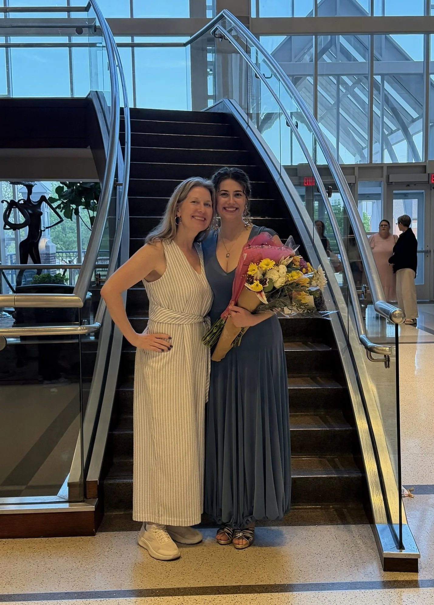 Two women standing in front of a staircase inside a modern building, smiling and holding a bouquet of flowers. One woman is wearing a striped dress and sneakers, the other in a long blue dress and sandals.