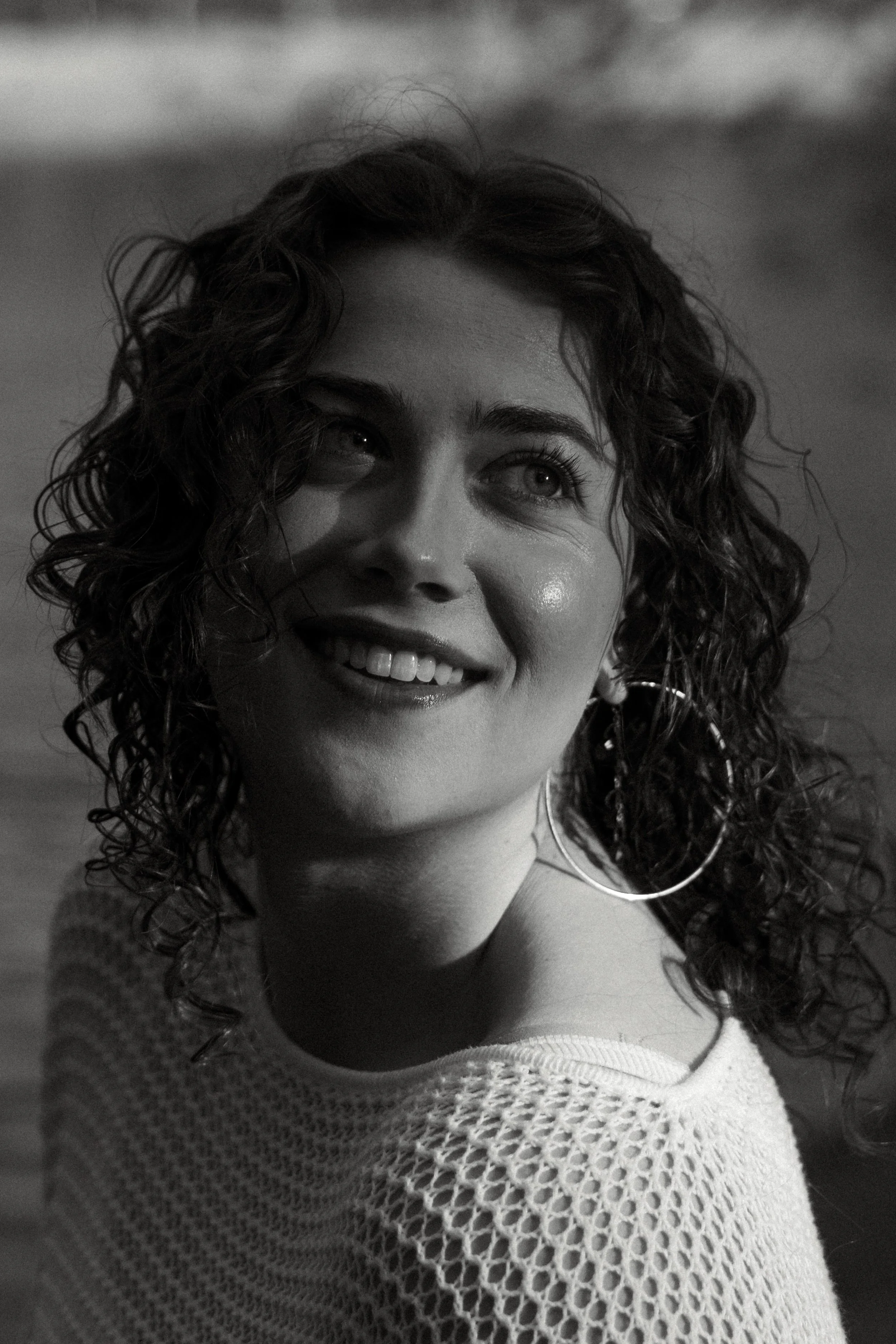 A woman with curly hair smiling, wearing hoop earrings and a patterned sweater, in a black and white photo.