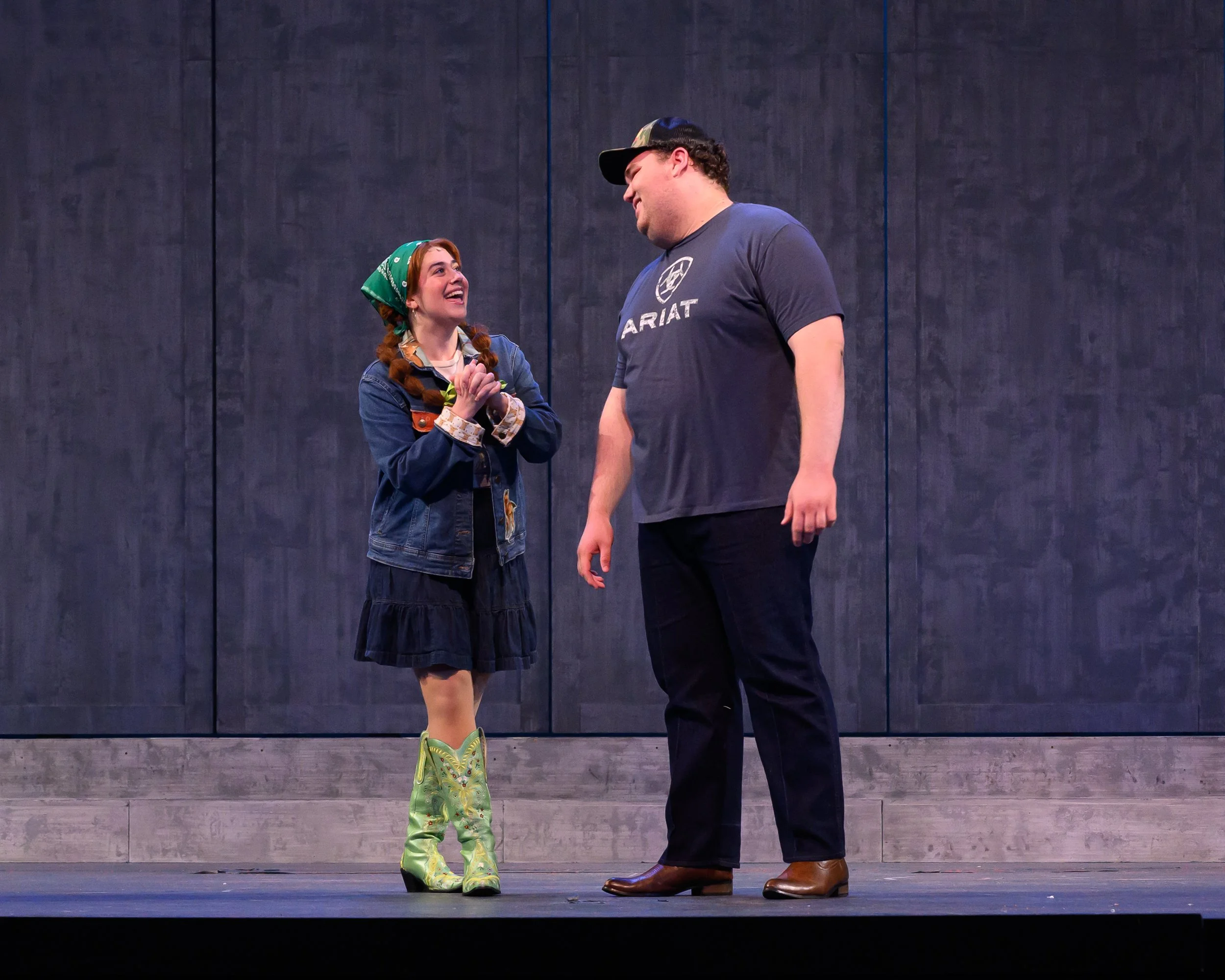 A woman with red hair, wearing a green bandana, denim jacket, and green cowboy boots, is smiling and holding hands with a man in a gray t-shirt, black pants, and brown shoes, standing on stage against a dark background.