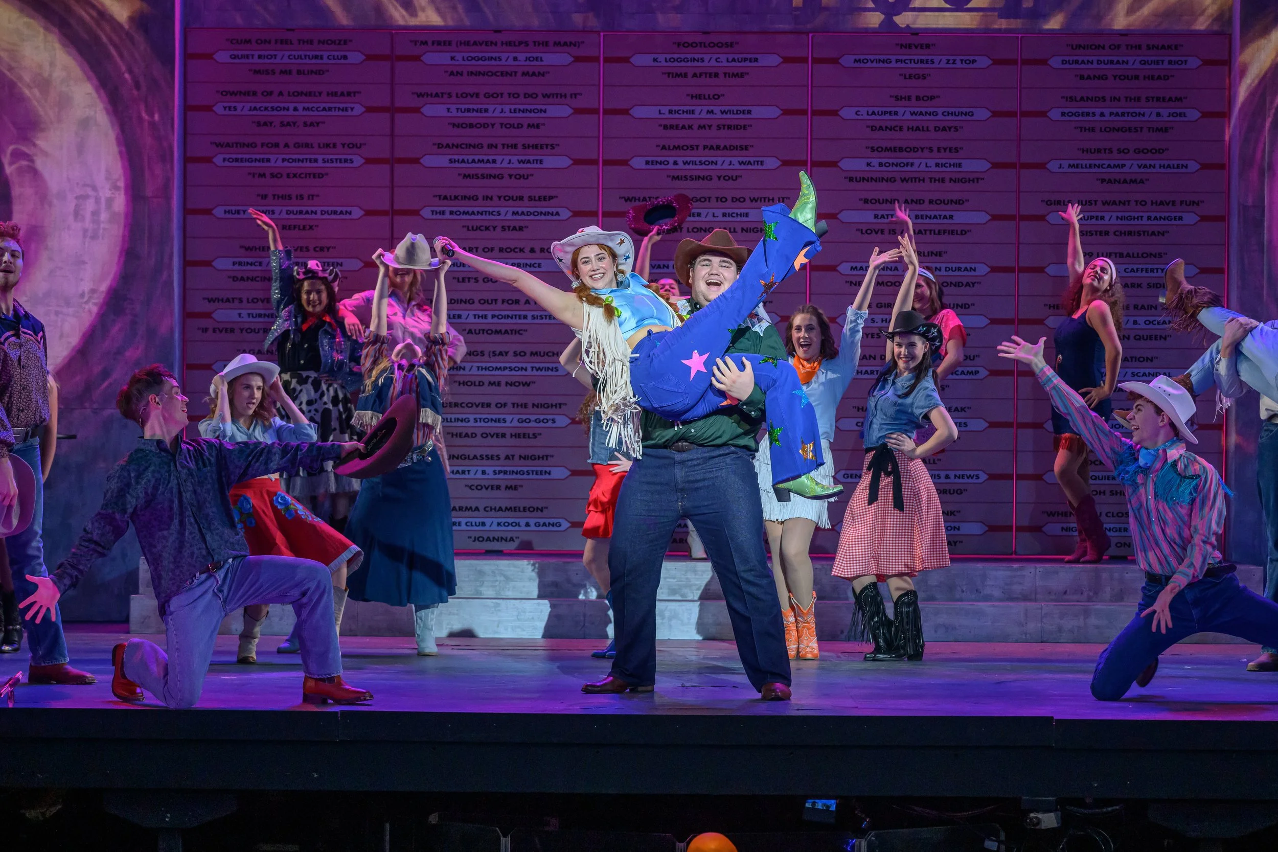 The cast of 'Oklahoma!' performing on stage, dressed in cowboy and western costumes, with a colorful backdrop featuring song titles.