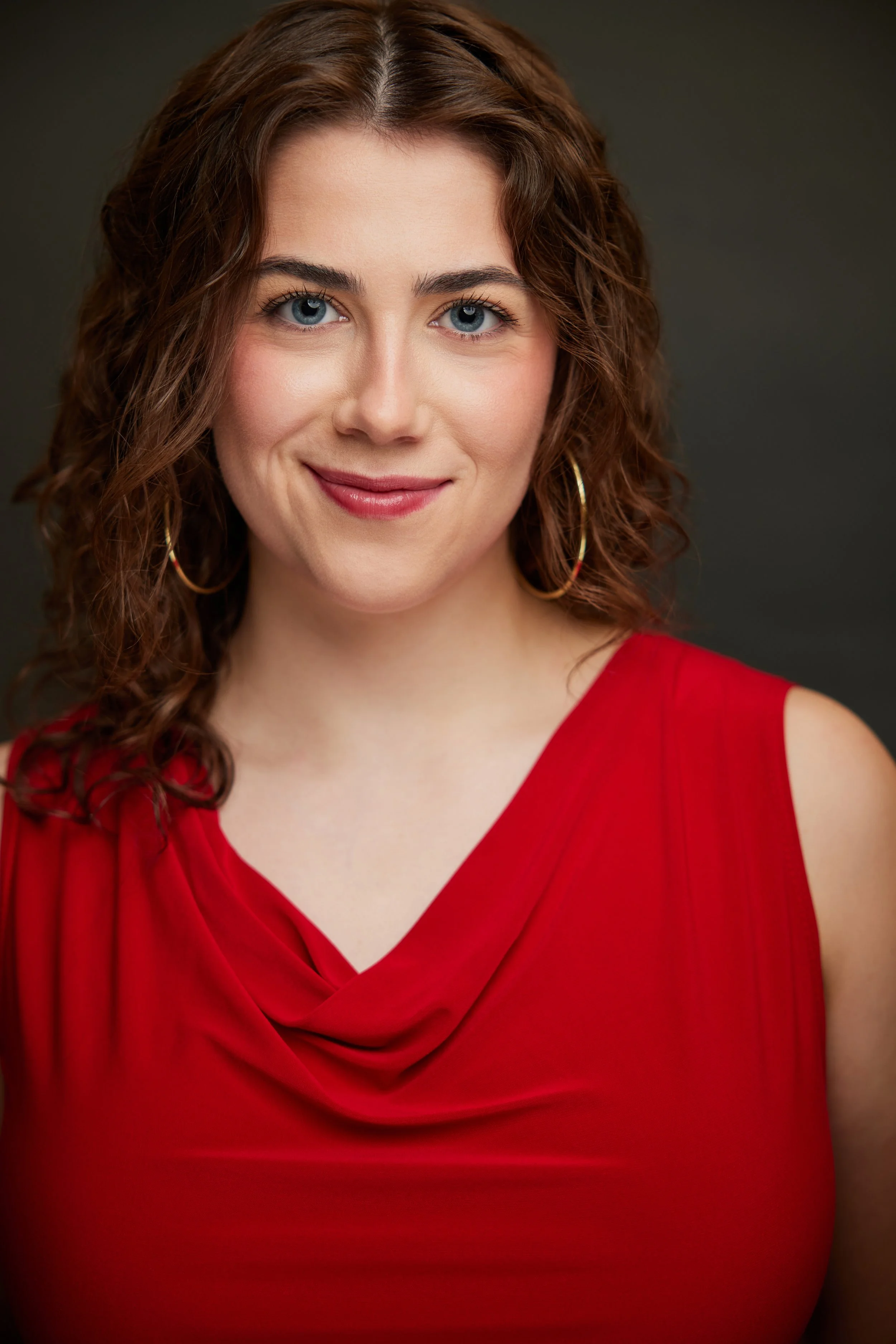 A woman with curly brown hair, blue eyes, and hoop earrings, wearing a red dress with a draped neckline, smiling softly against a dark background.