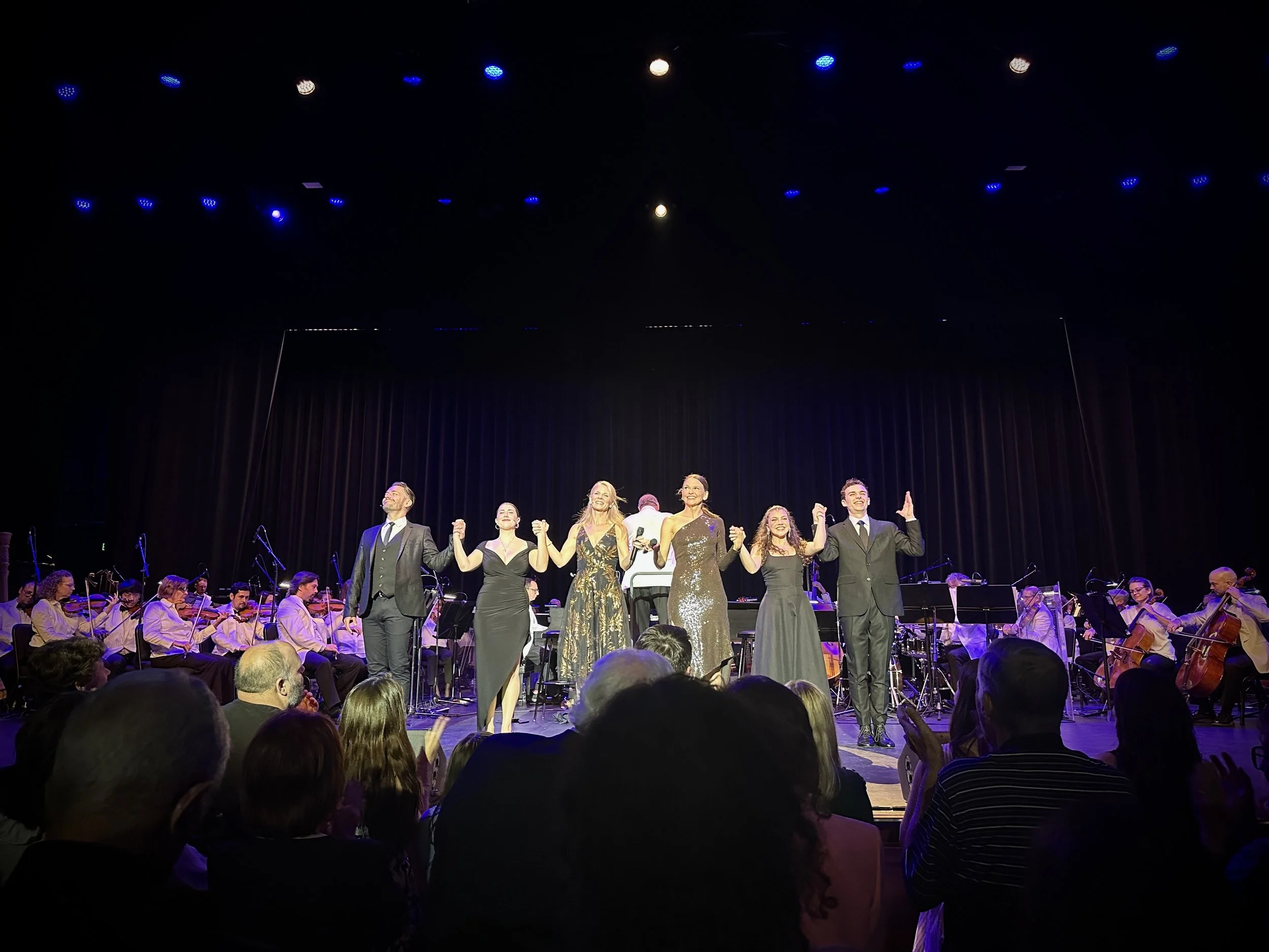 The image shows a group of performers on stage, holding hands and taking a bow after a performance. Behind them, there is an orchestra with musicians playing string instruments. The stage is lit with blue and white lights, and the audience is visible