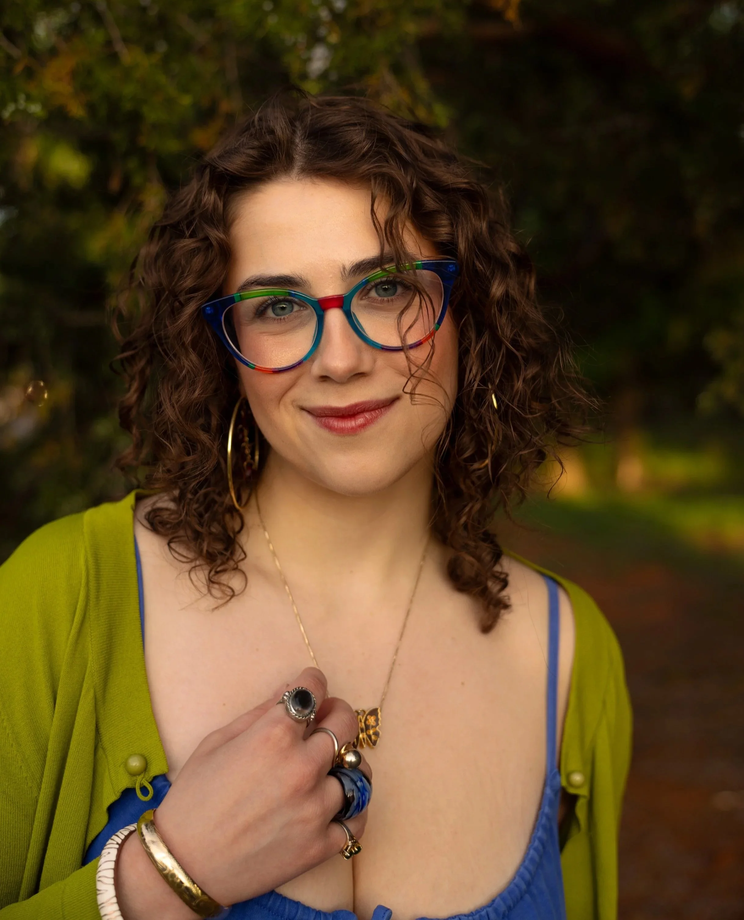 Close-up photo of a woman with curly brown hair, wearing colorful glasses, jewelry, and a blue top with a green jacket, outdoors with blurred green foliage background.