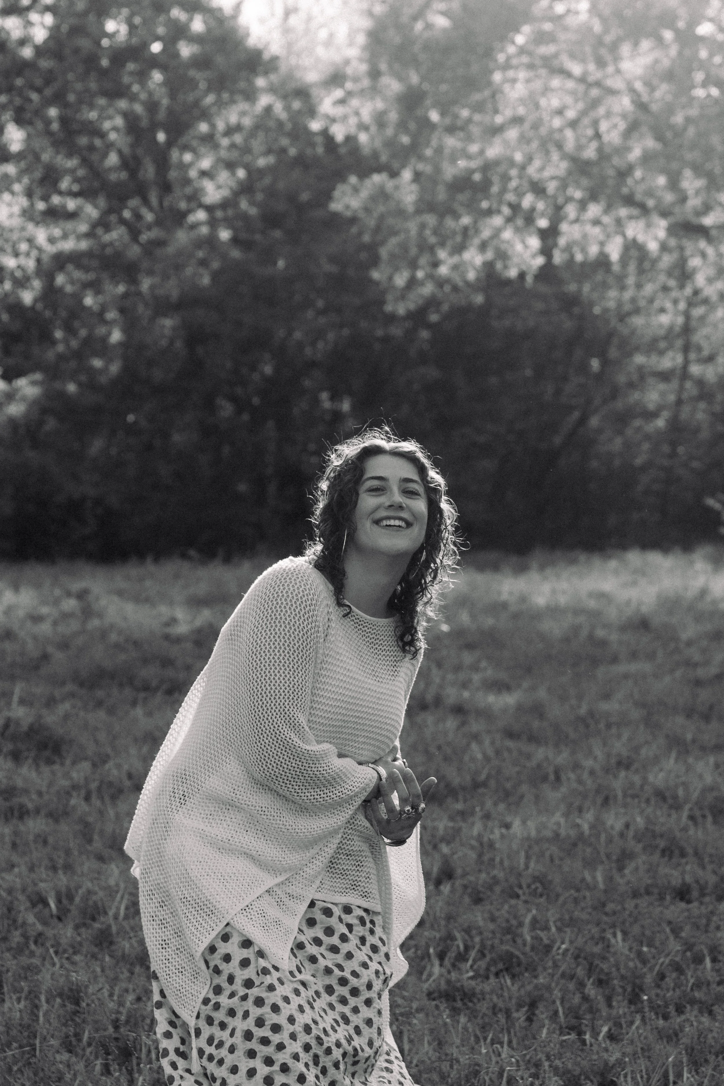 A young woman with curly hair smiling outdoors, wearing a light sweater over a polka dot dress, standing in a grassy field with trees in the background.