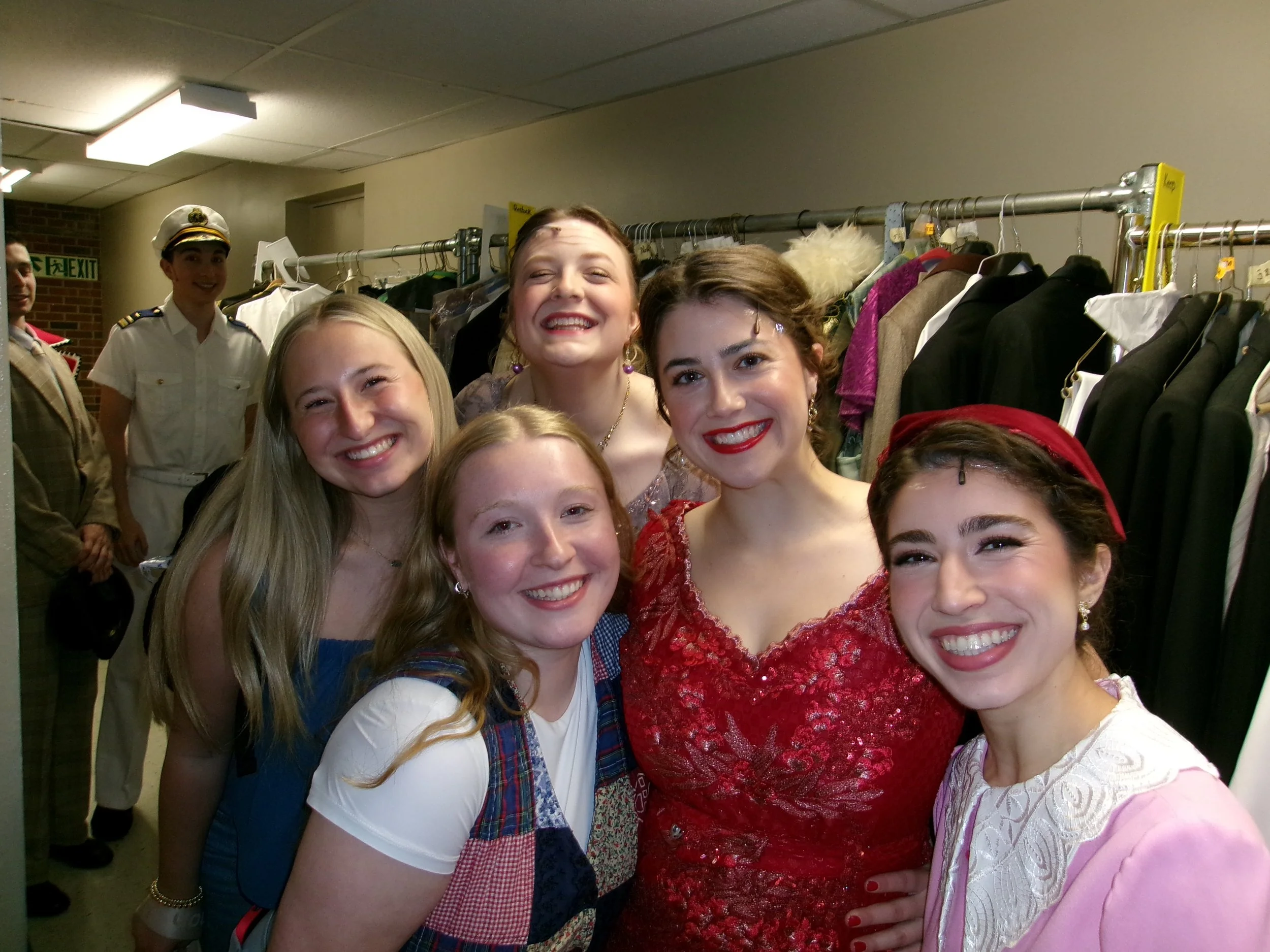 Group of women dressed in colorful costumes, smiling and taking a selfie in a backstage area with racks of clothing in the background.
