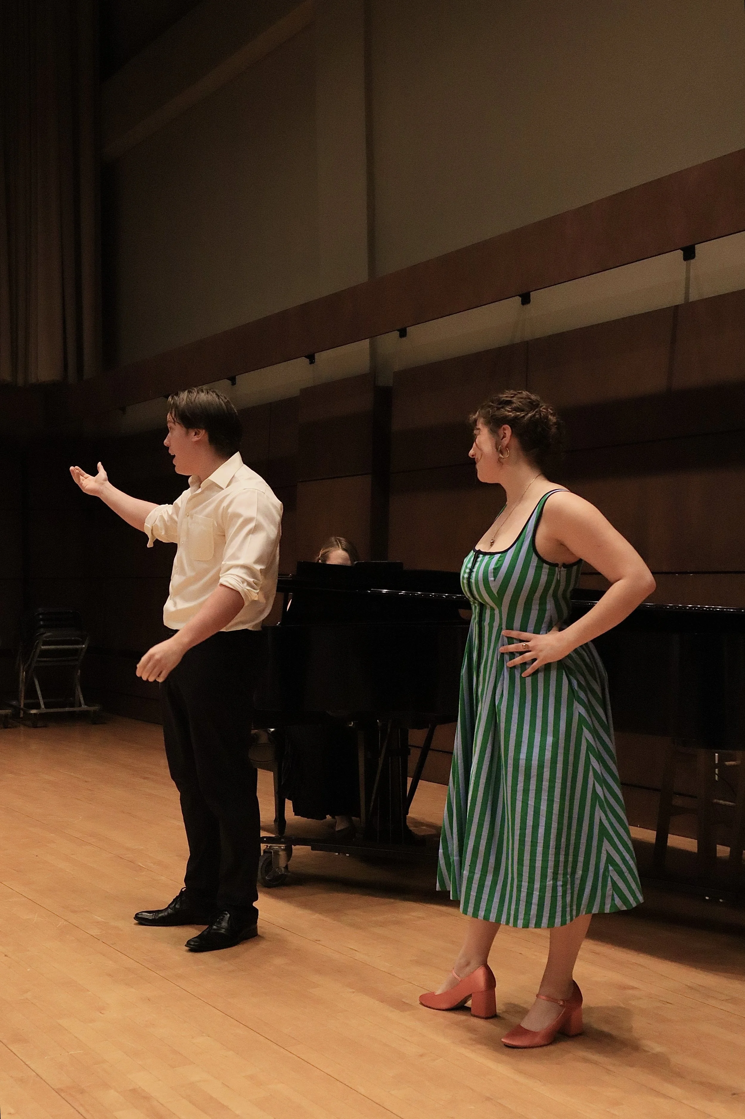 A man and a woman are performing on a stage in a music hall. The man is dressed in a white shirt and black pants, and is gesturing with his hand. The woman is wearing a green and white striped dress with pink high heels, and is standing with her hand