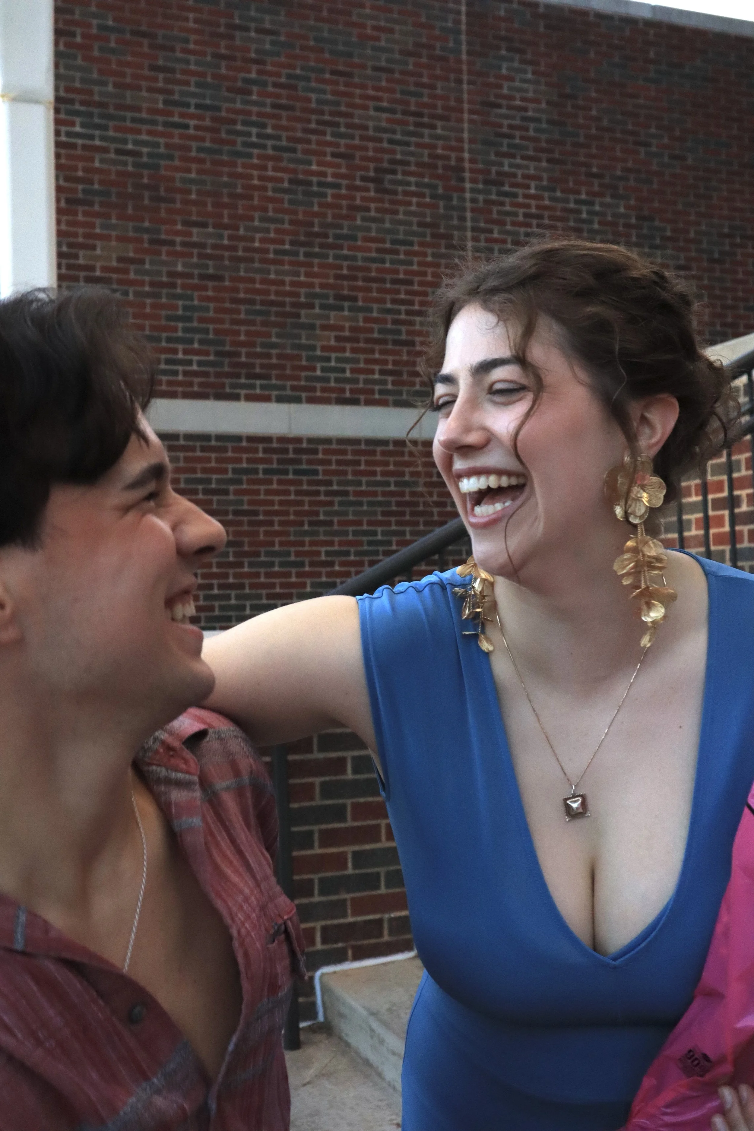 A woman with short dark hair and hoop earrings is smiling and laughing with a man with dark hair and light skin, who is wearing a plaid shirt. They are standing outside, in front of a brick wall, and appear to be enjoying a joyful moment together.