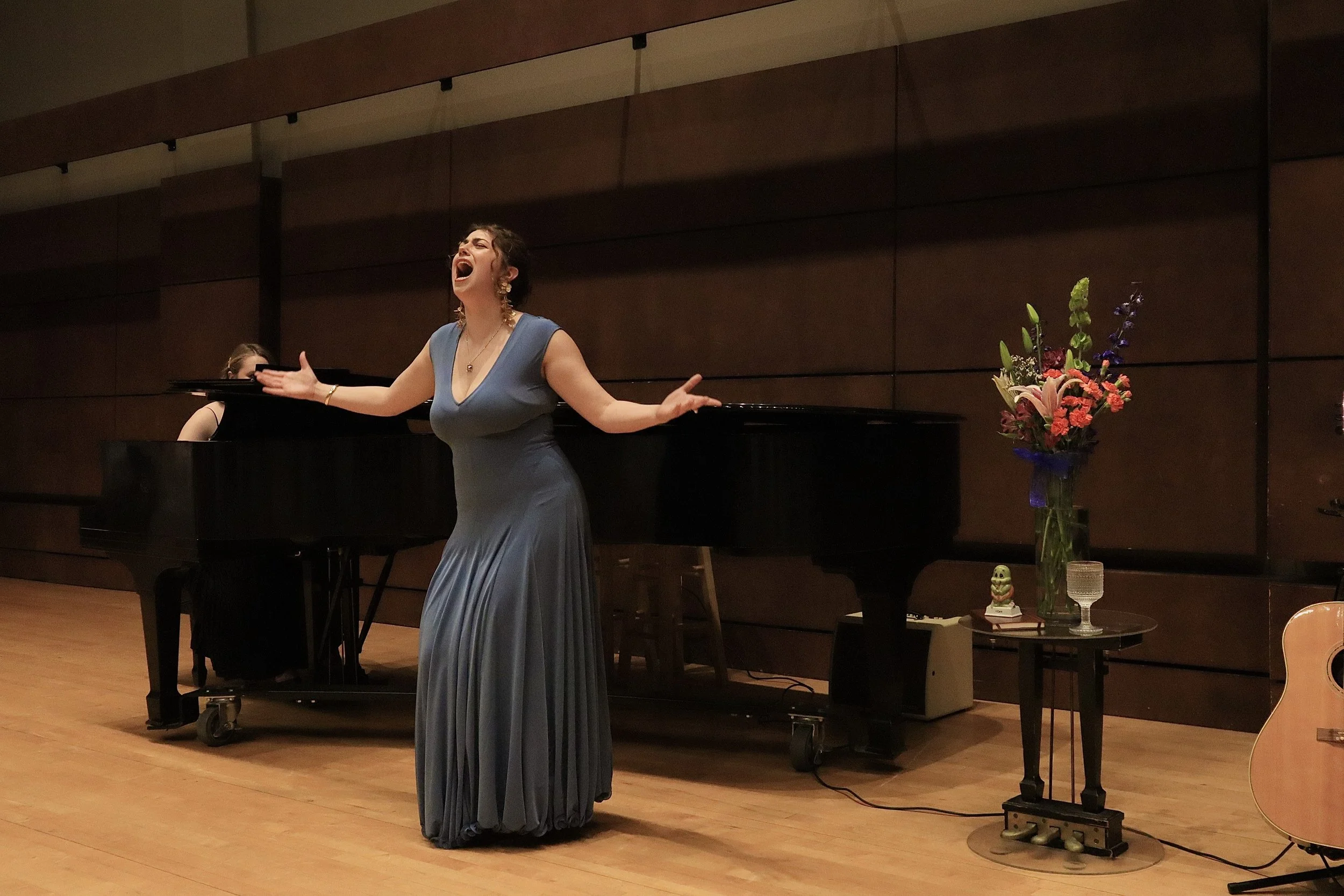 A woman singing passionately with her arms outstretched, standing near a pianist in a performance room. A flower arrangement and other decorative items are on a table nearby.