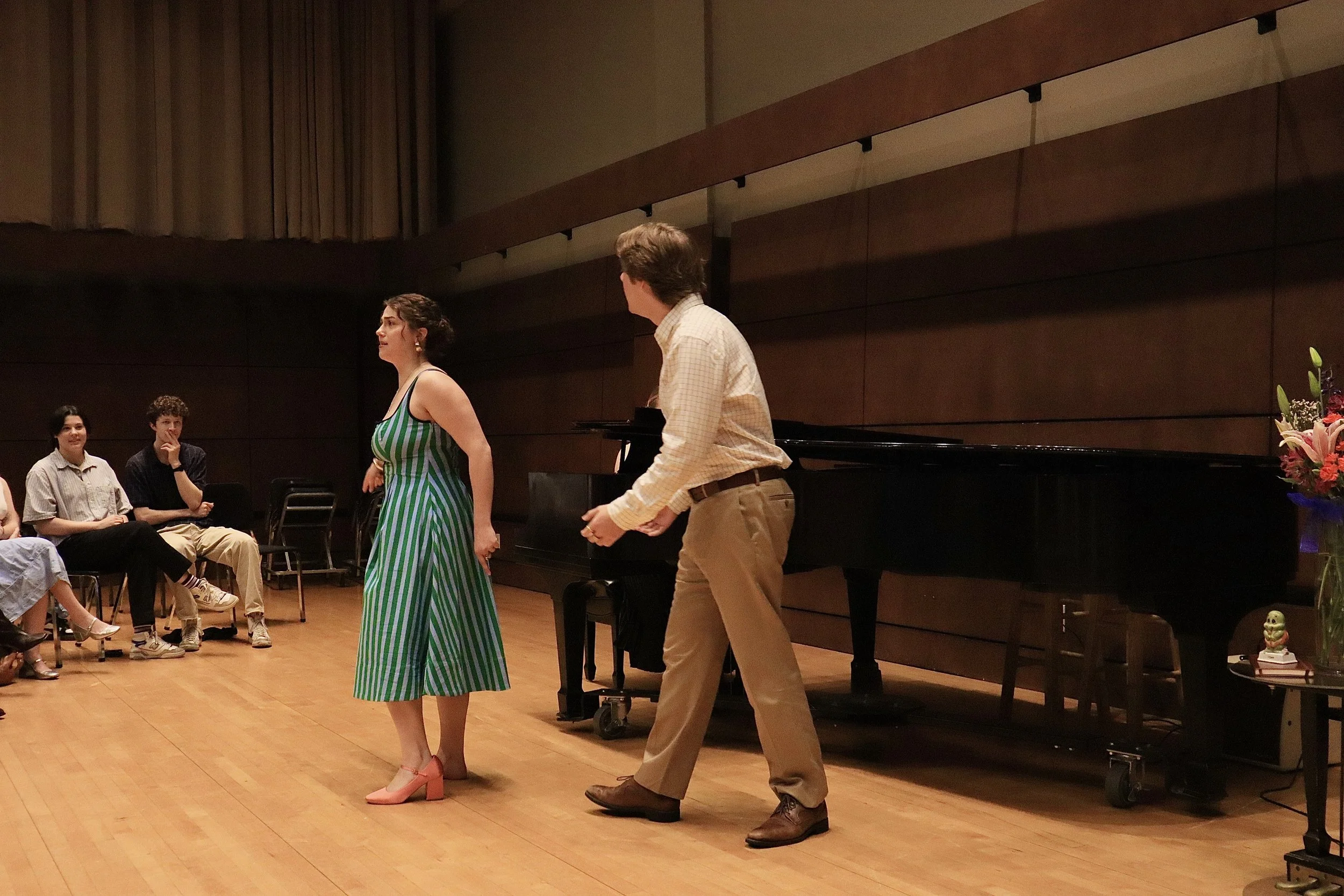 A group of people gathered on a stage in a concert hall or auditorium. Two people are standing at the center, a woman in a green and white striped dress and a man in a checkered shirt and tan pants. Other individuals are seated and watching. There is